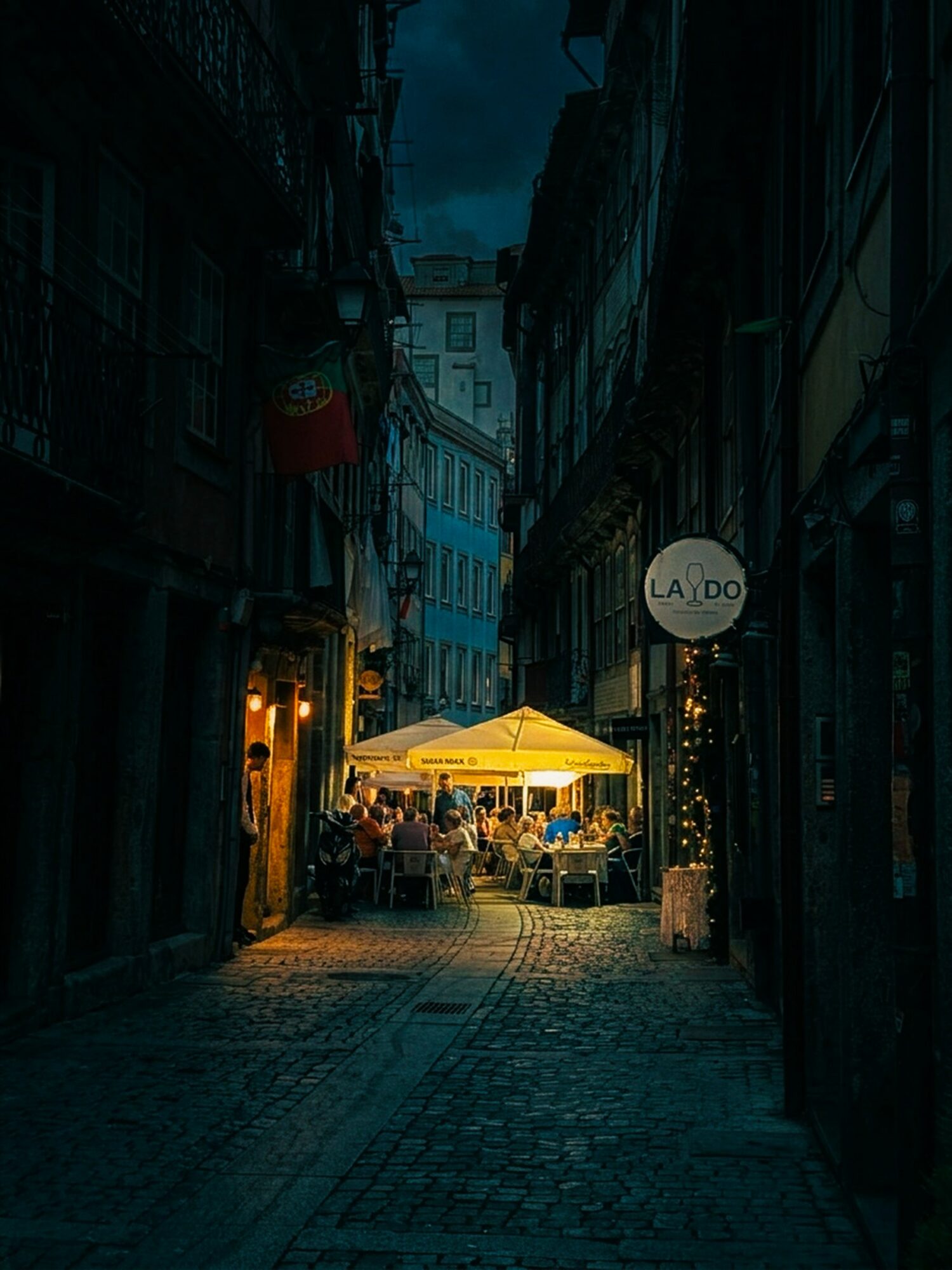 Night scene of a narrow street with outdoor seating under a yellow umbrella, illuminated by warm lights, buildings on both sides.