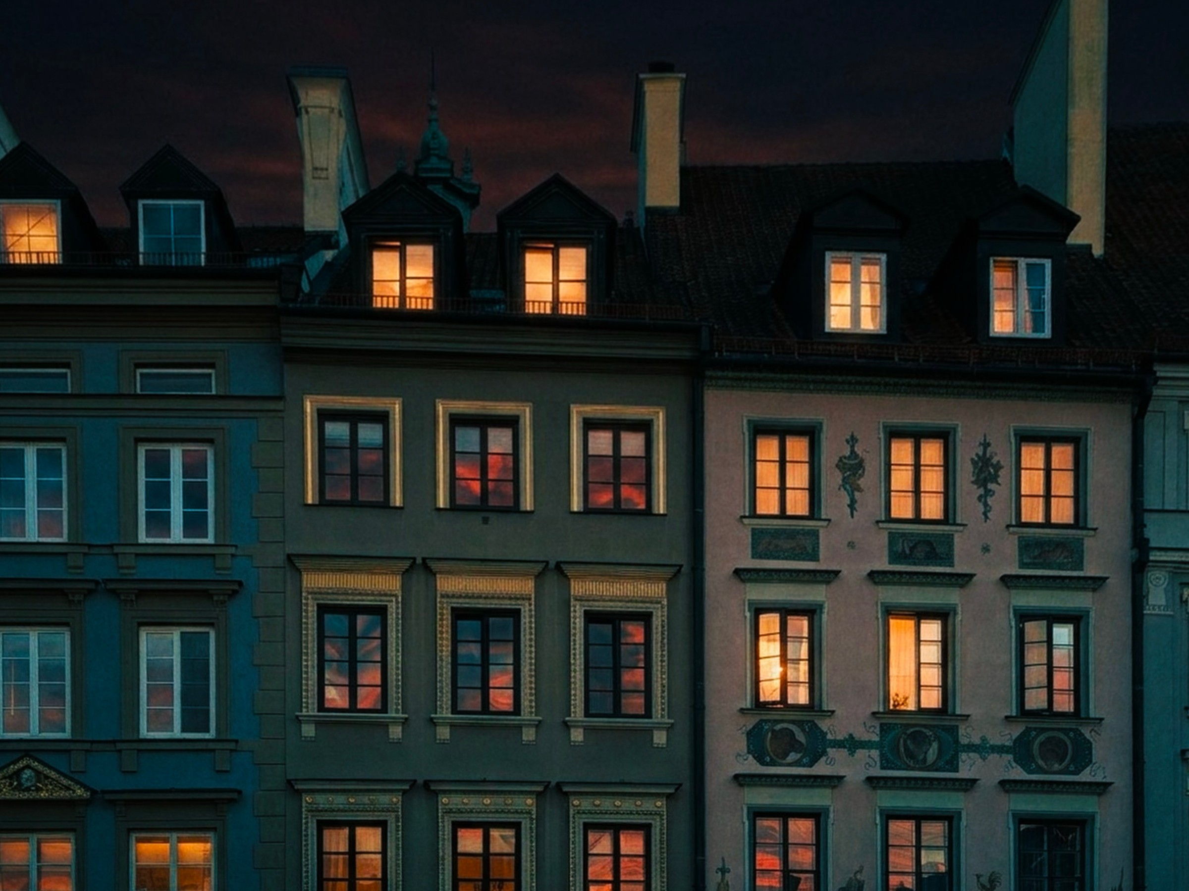Nighttime view of a row of buildings with illuminated windows, dark sky, and chimneys on rooftops.