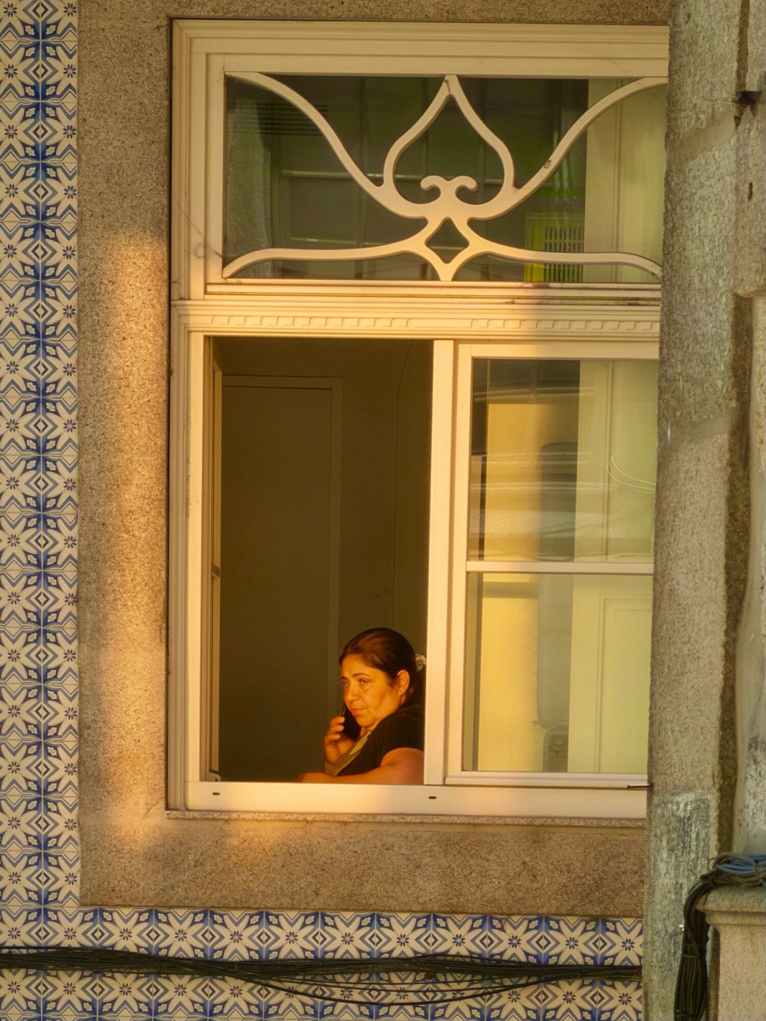 Woman looking out of a window with decorative glass design, sunlight illuminating her face, building exterior visible.