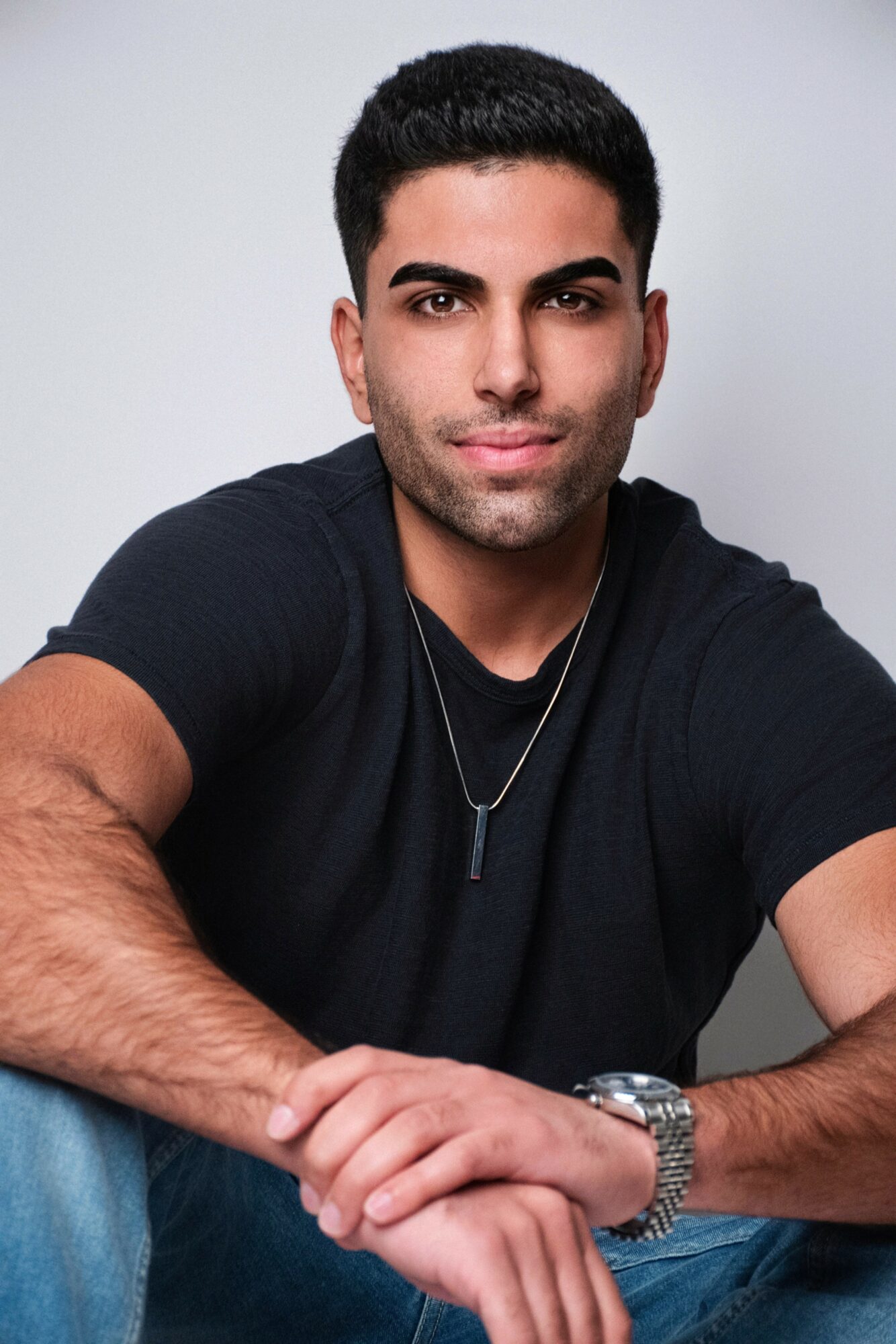 Young man with short dark hair and a slight smile, wearing a black t-shirt, sitting with arms crossed, against a plain light background.
