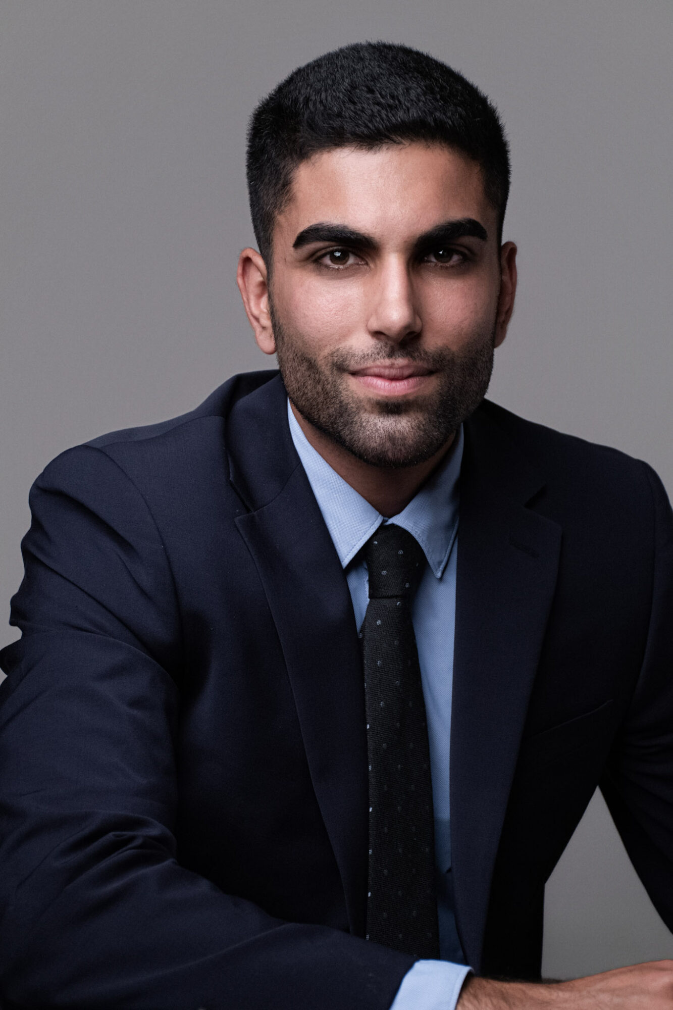 Portrait of a young man with dark hair and beard, wearing a dark suit, light blue shirt, and dark tie, against a gray background.