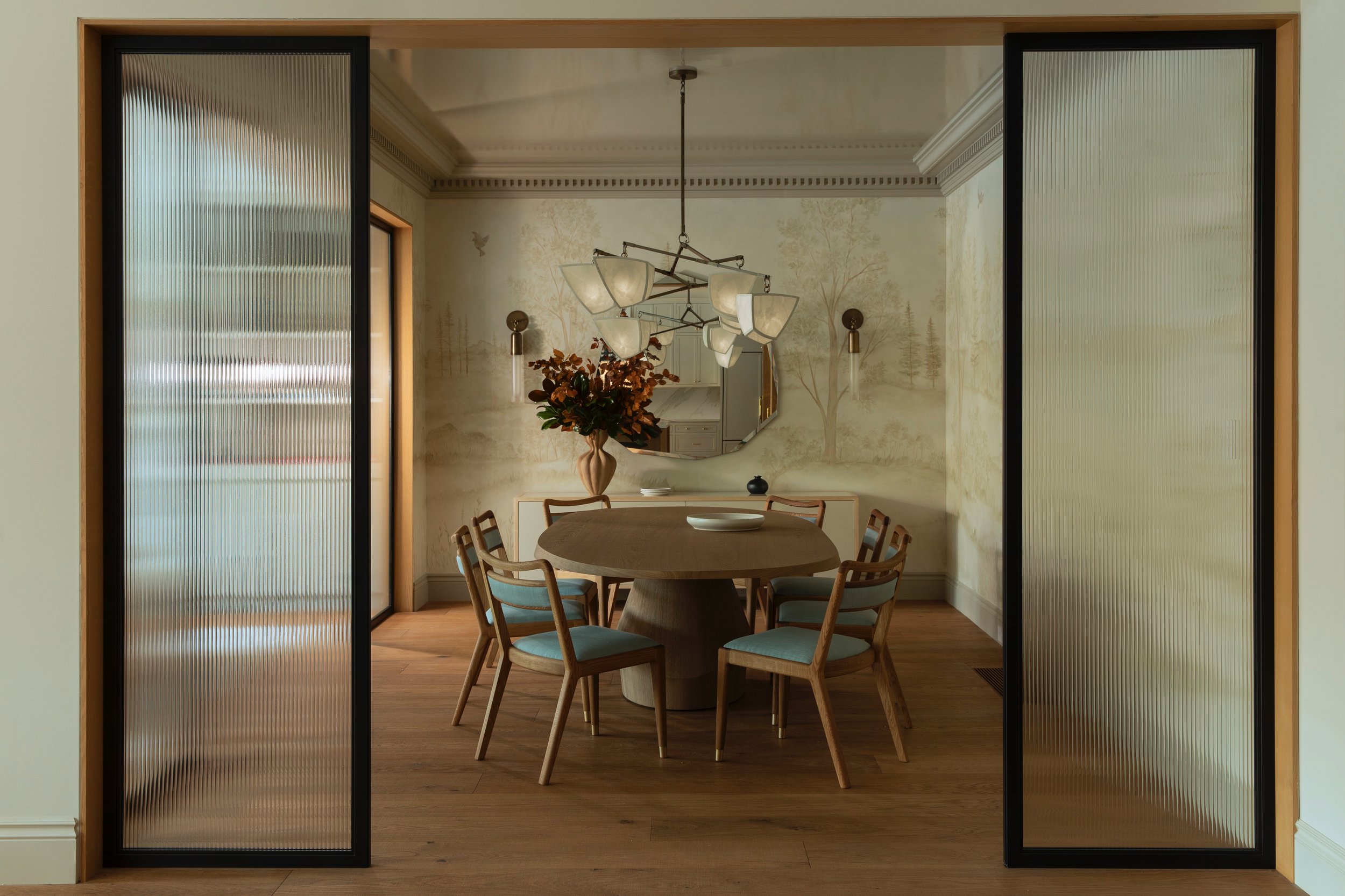 Dining room with round table, six chairs, chandelier, and a vase with flowers, viewed through sliding glass doors.