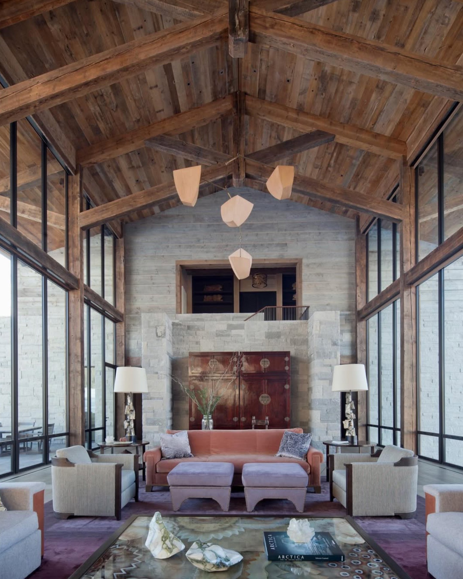 Living room with high wooden ceiling, large windows, and modern furniture including sofas, armchairs, and lamps, with a decorative wall piece.