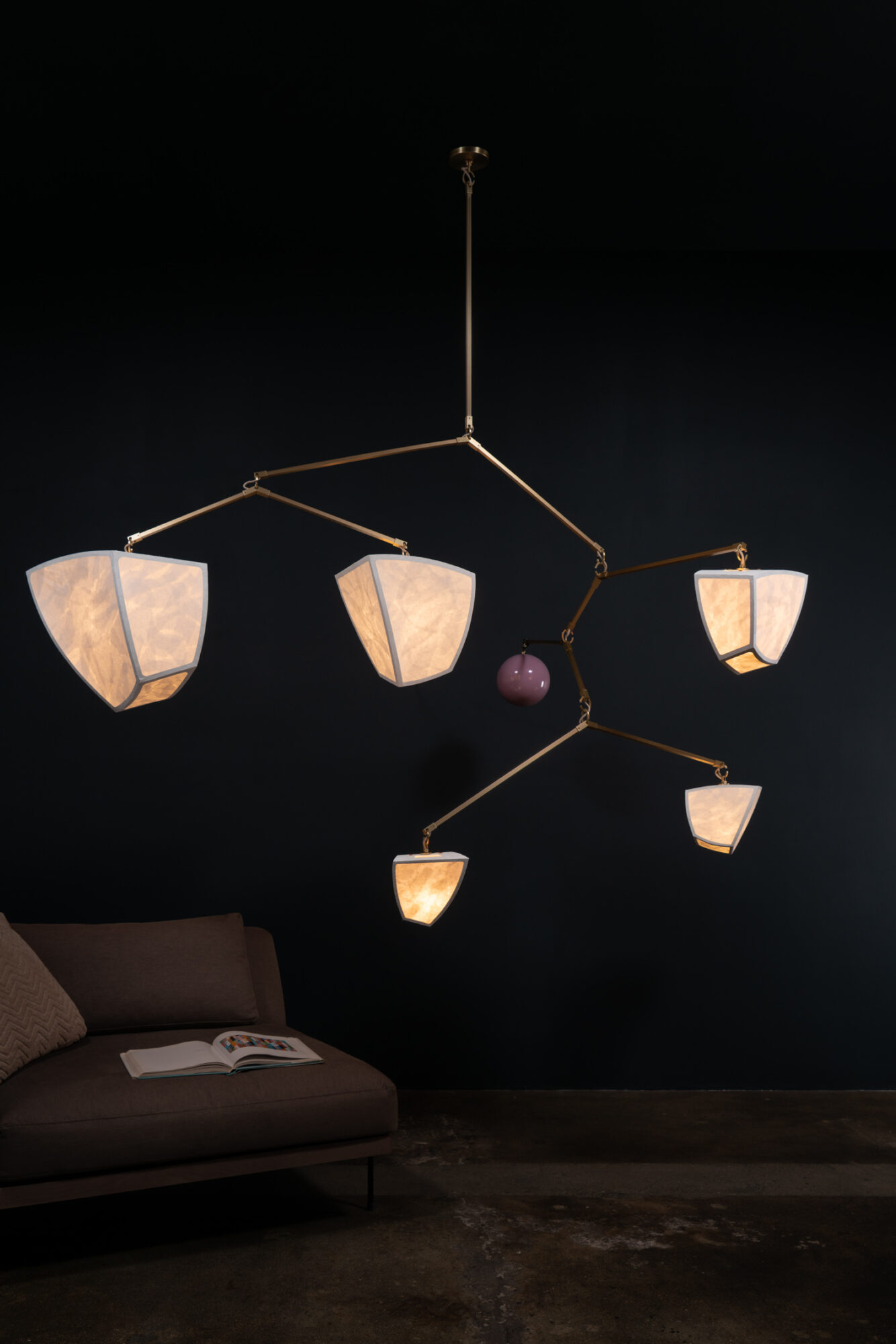 Modern chandelier with five lampshades hanging from a dark ceiling, with a sofa and book below, against a dark wall.