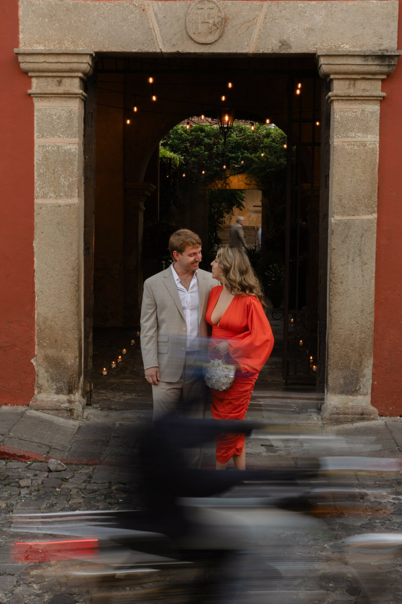 Two people stand in a doorway, one in a light suit and the other in a red dress, with a tree visible inside.