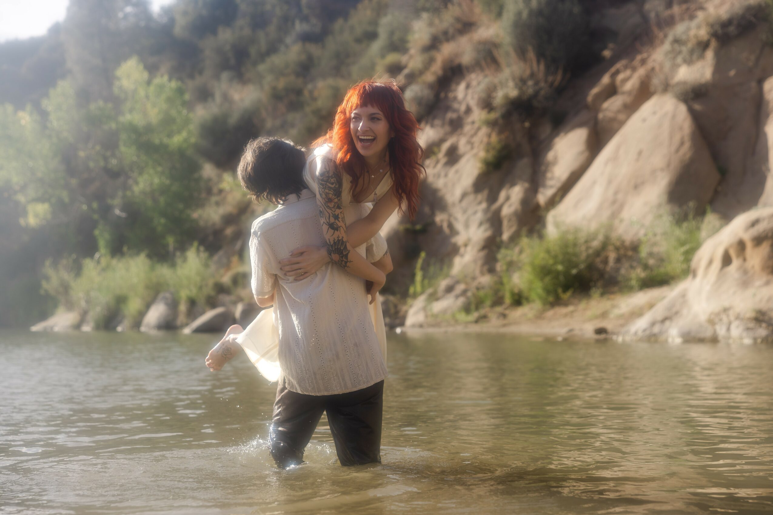 Woman holding a child in water near a rocky shoreline with trees and rocks in the background.