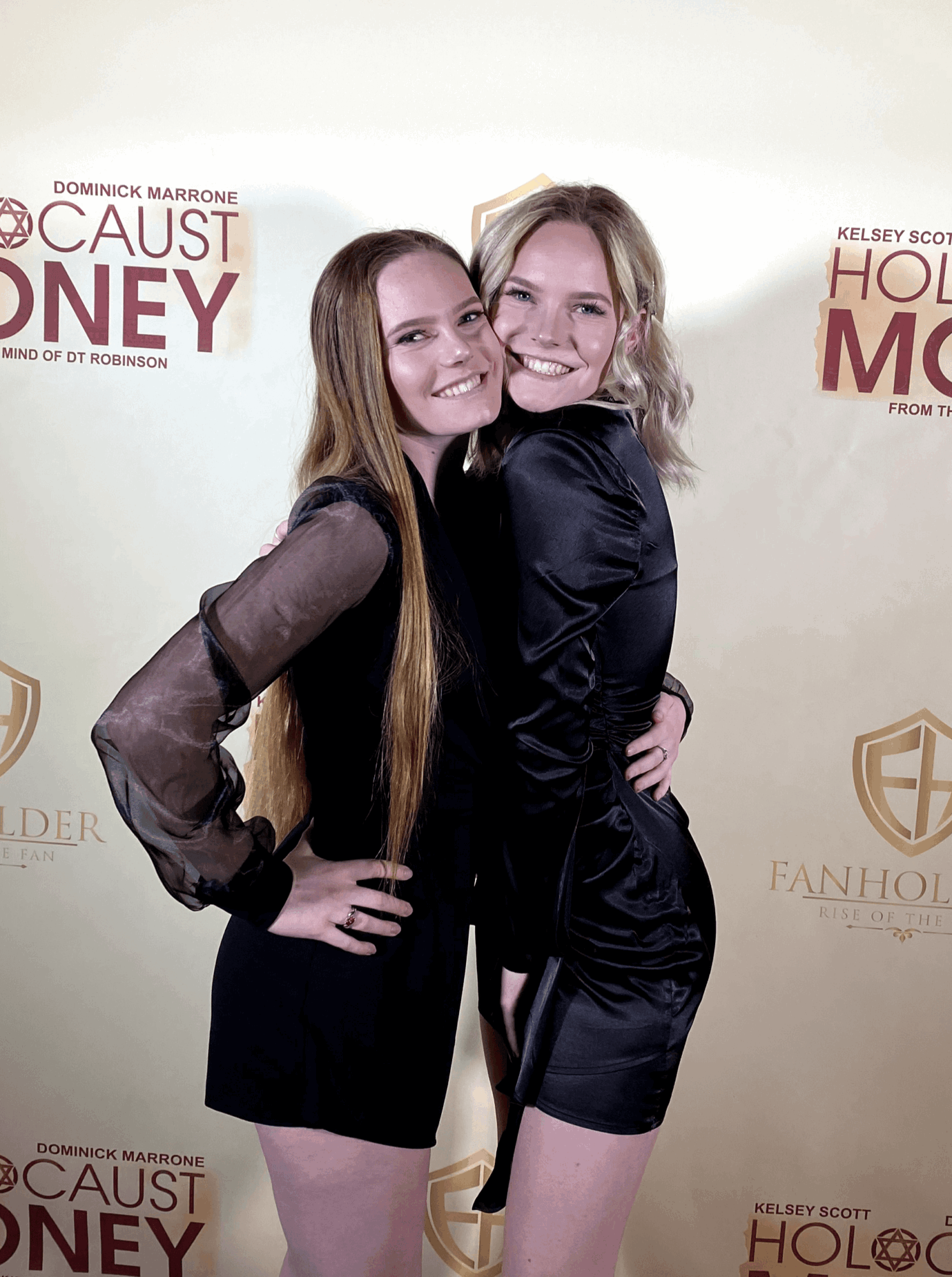 Two young women smiling and hugging in front of a backdrop with text and logos.