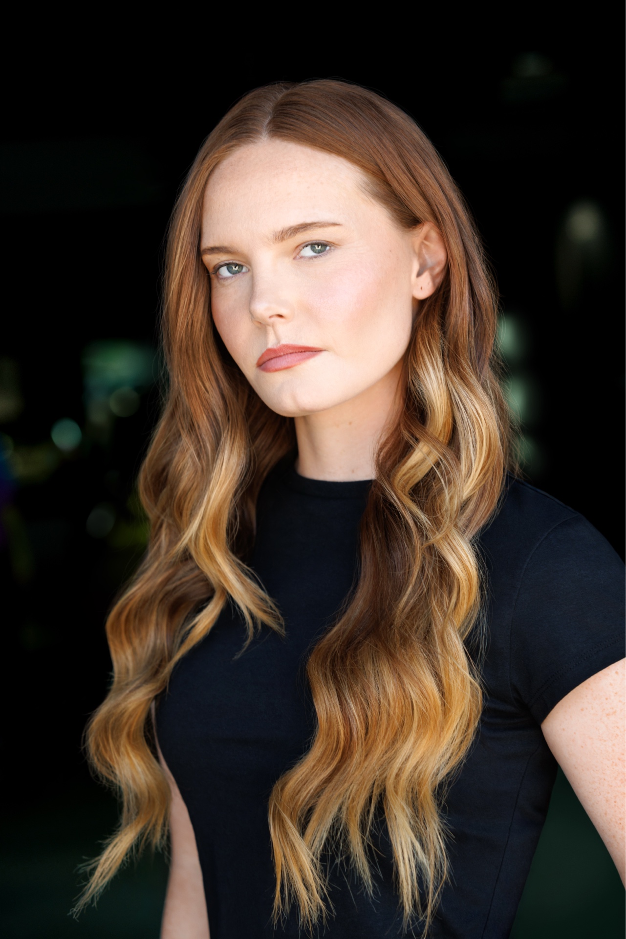 Young woman with long wavy hair and fair skin, wearing a dark top, looking at the camera against a dark background.