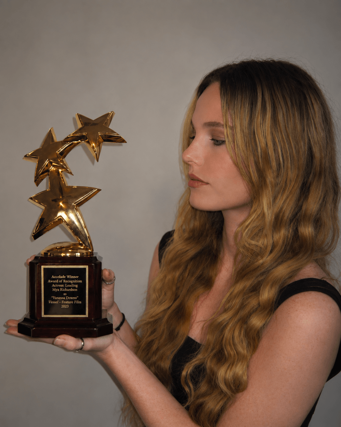 Woman with long wavy hair holding a trophy with three gold stars, looking at it, against a plain background.