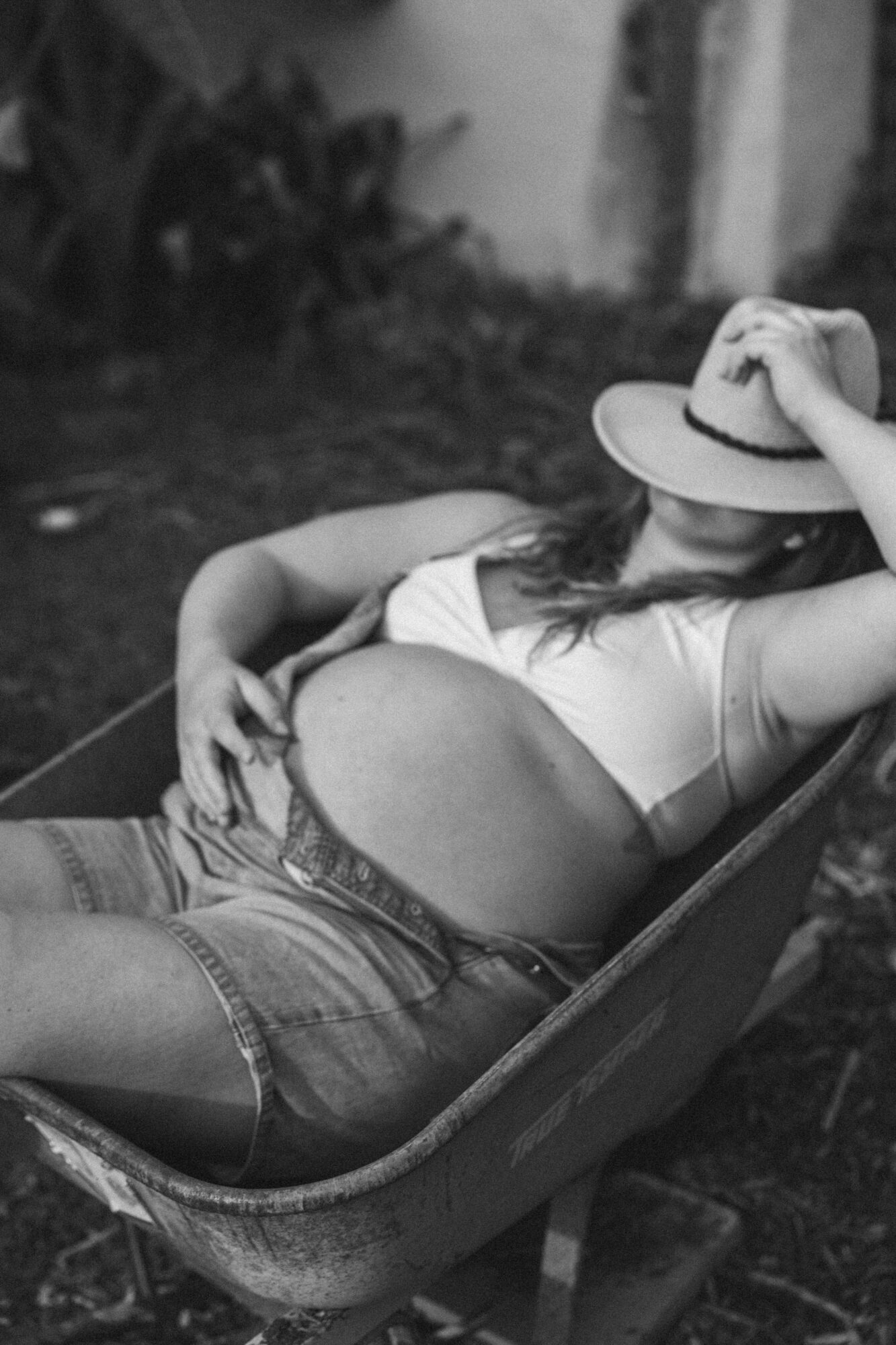 Pregnant woman reclining outdoors, wearing a hat and casual clothing, resting in a chair with her hand on her belly.