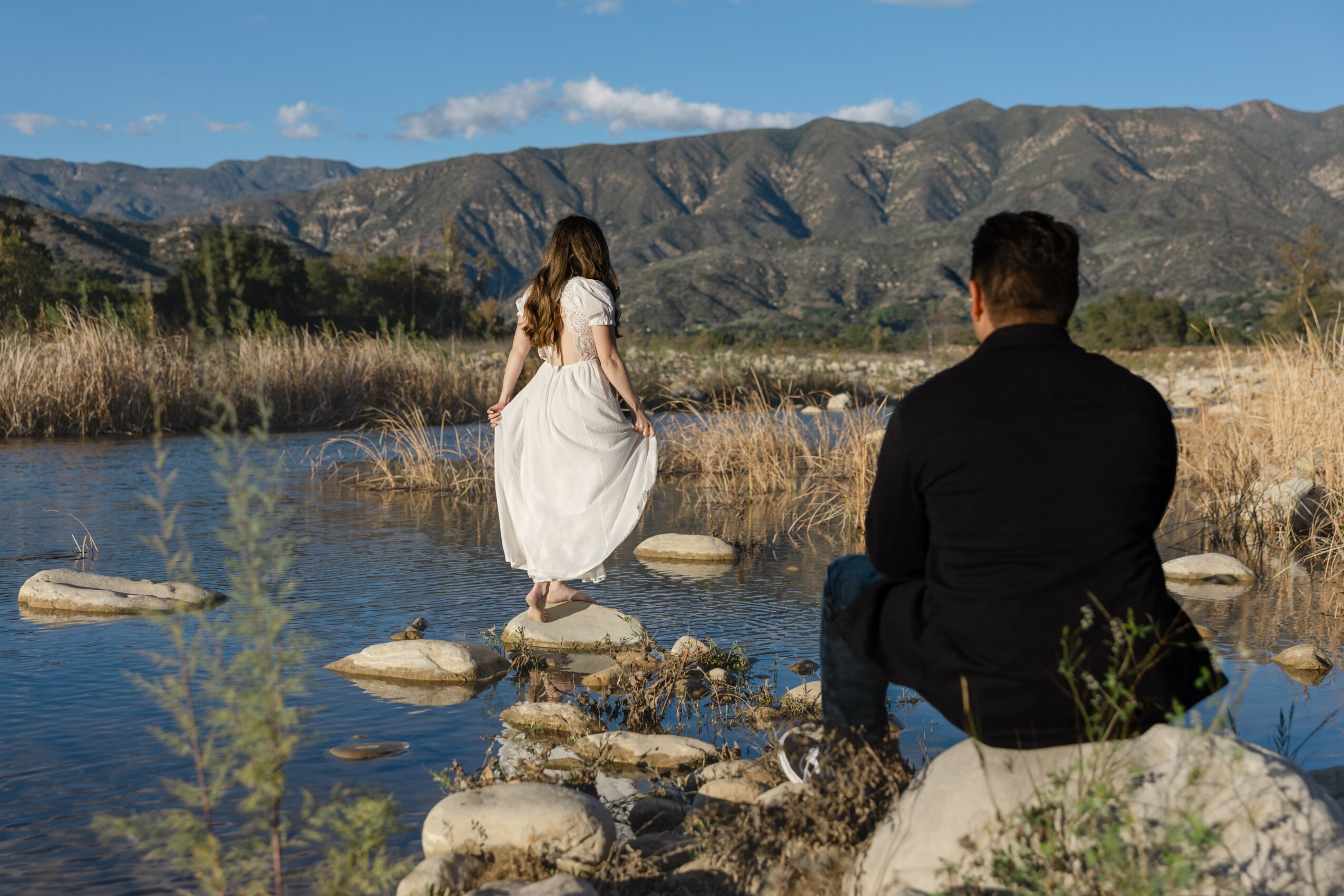 A woman in a white dress stands on rocks in a river, with mountains in the background and a man sitting nearby.