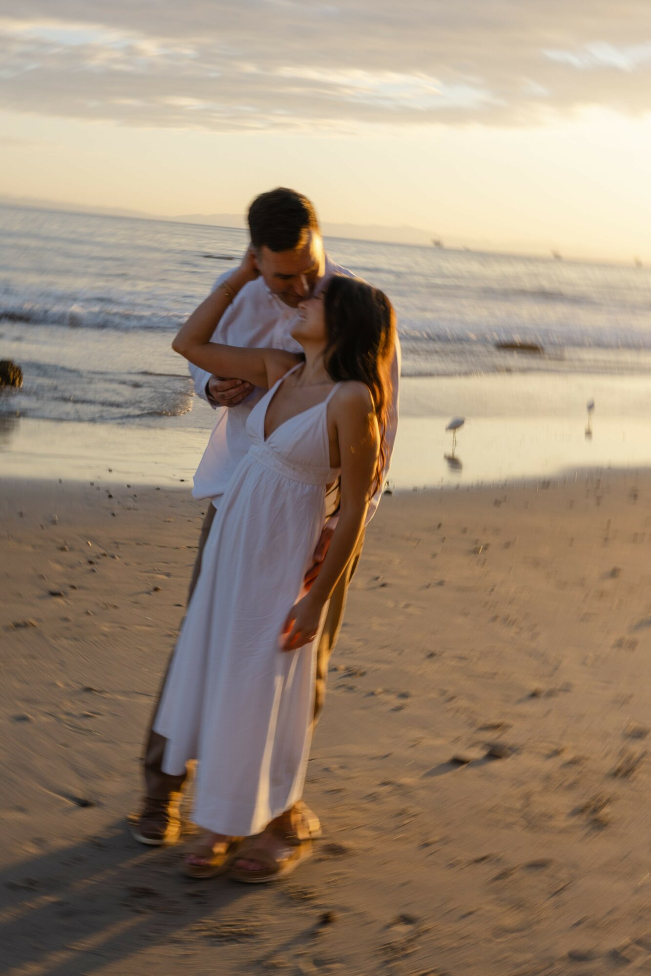 Two women in white dresses standing on a beach at sunset, embracing each other, with the ocean and sky in the background.