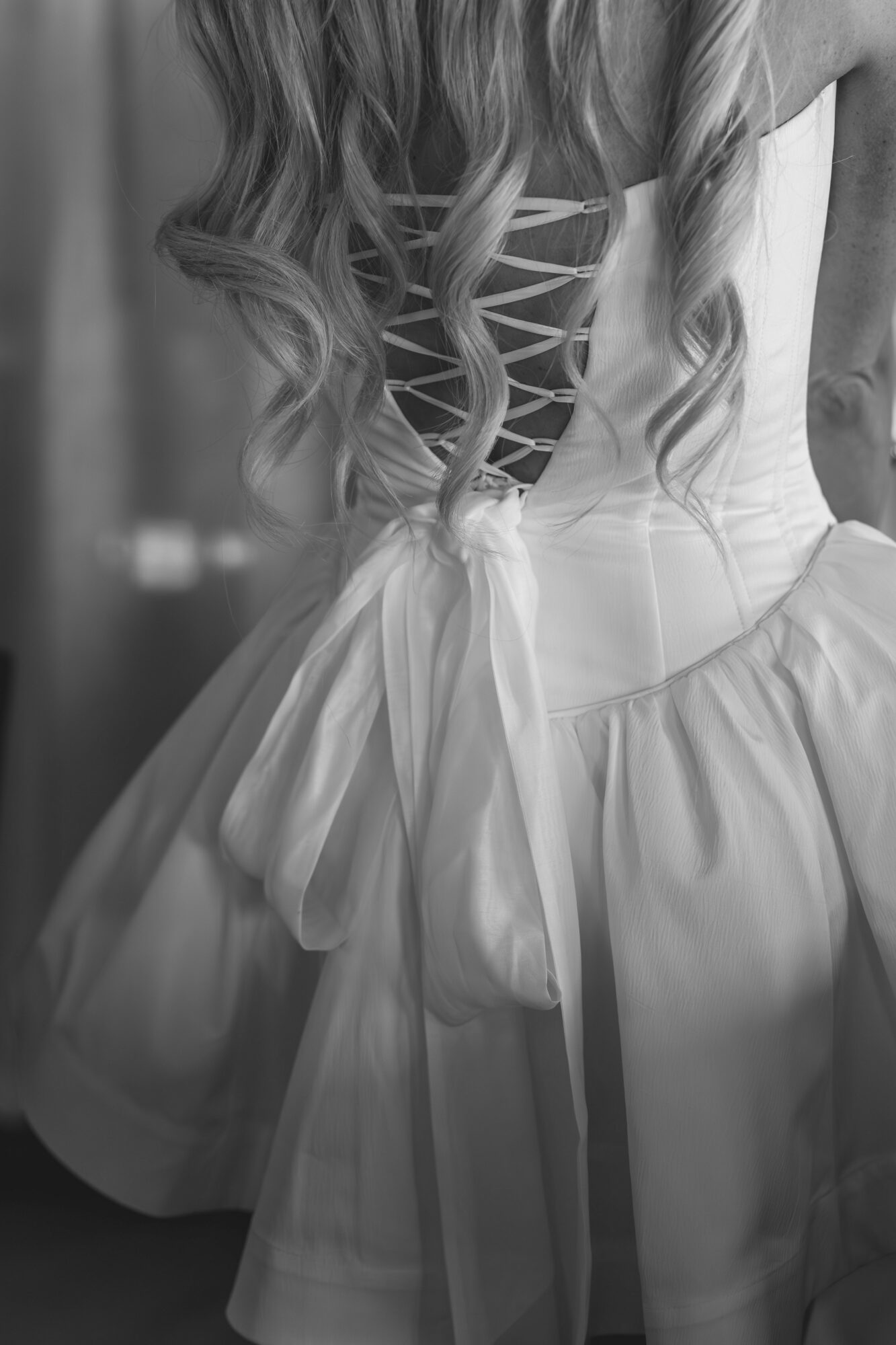 Back view of a person in a dress with a lace-up back and long, wavy hair, tied with a bow at the waist.
