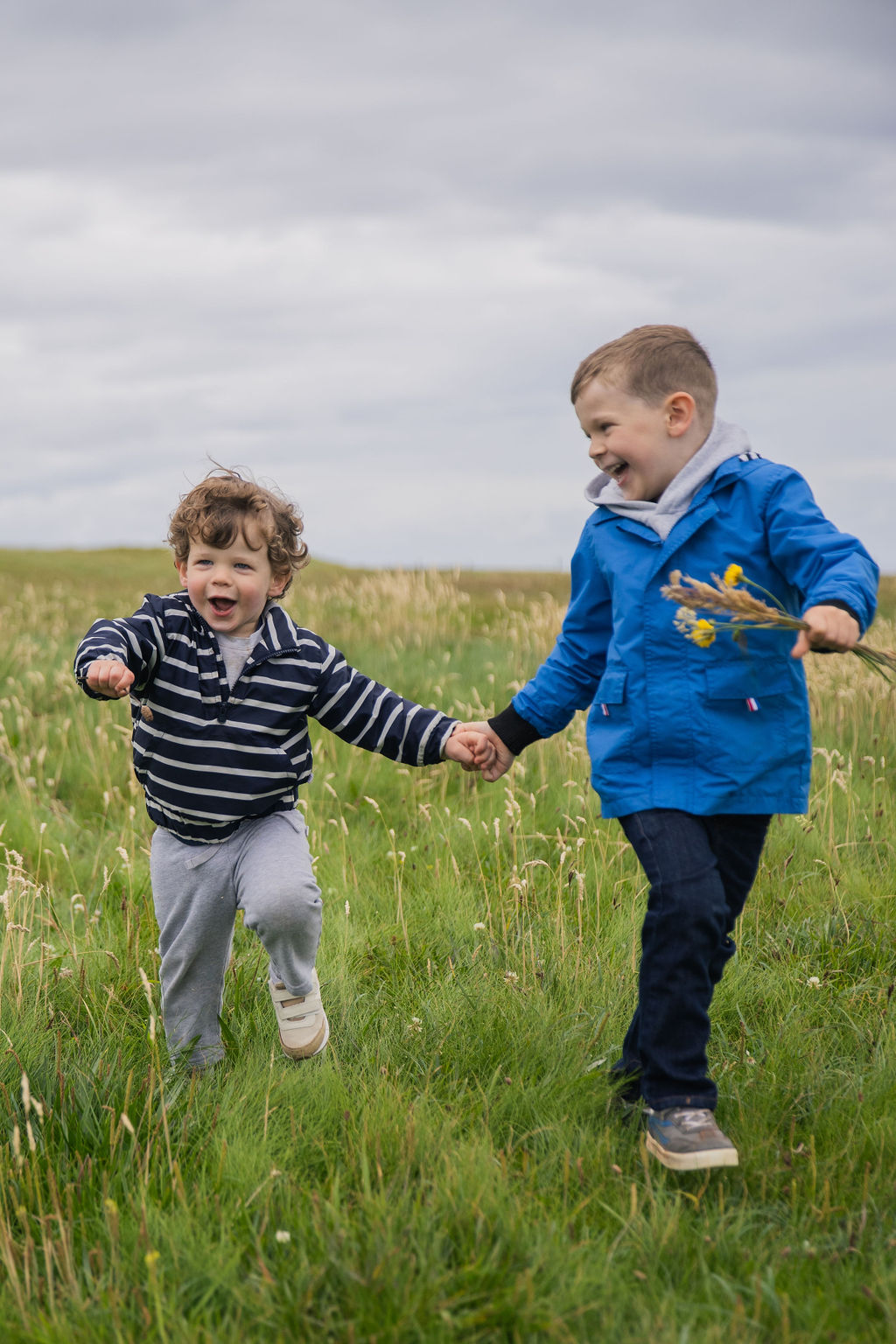 Two children holding hands and running in a grassy field under a cloudy sky, smiling and playing.