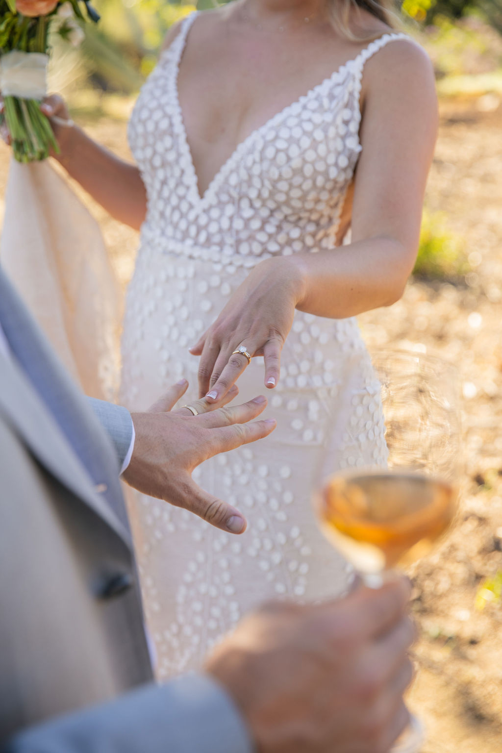 Close-up of a woman in a white dress with textured polka dots, holding hands with a person in a suit, outdoors.