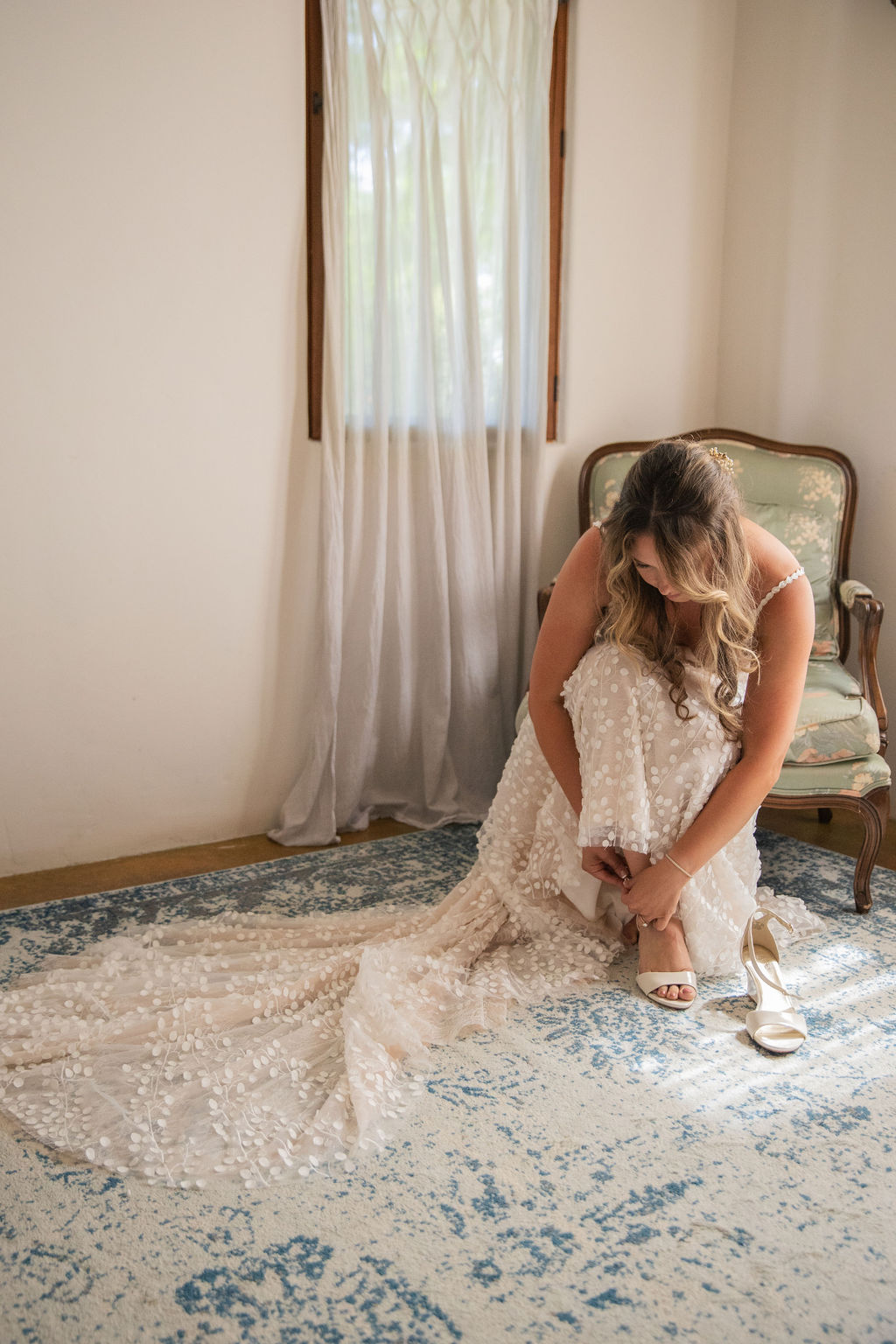 Woman in a wedding dress sitting on a vintage sofa, adjusting her shoe in a bright room with a window and curtain.