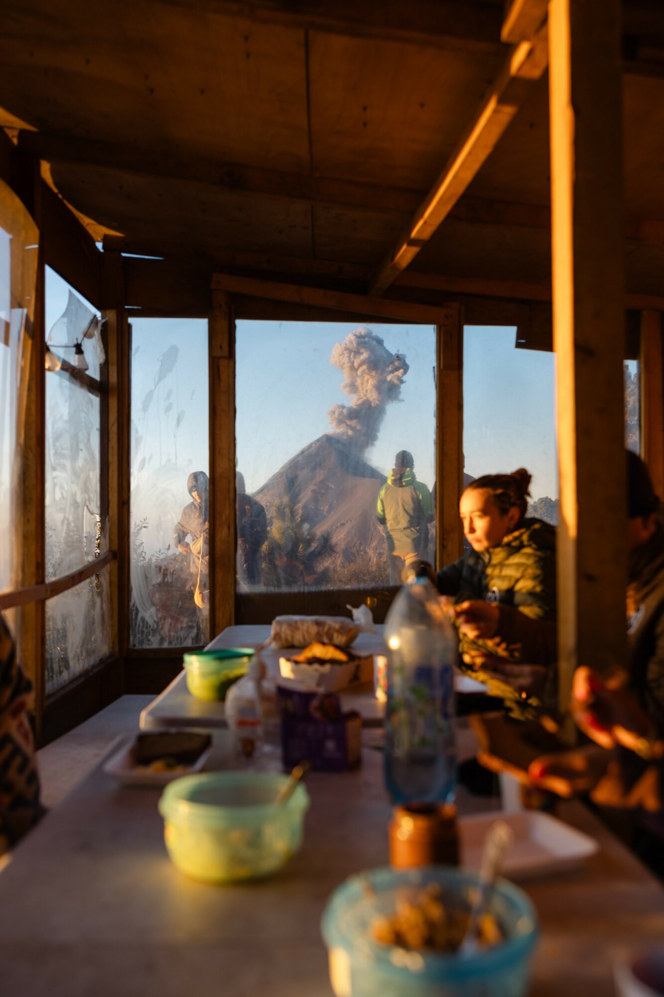 People sitting at a table inside a wooden structure with large windows, volcano erupting in the background, daytime scene.