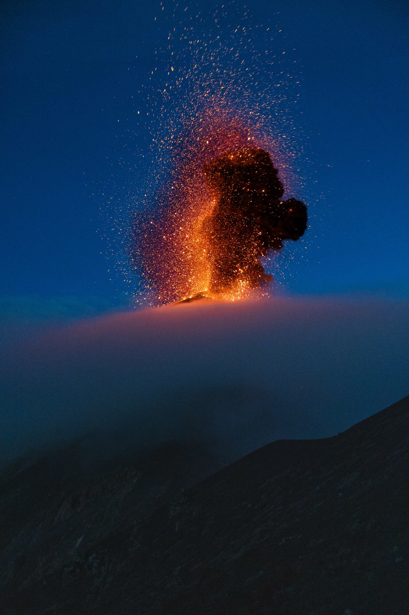 Eruption of volcanic ash and lava against a dark sky, with glowing orange and red colors, and a cloud of smoke.
