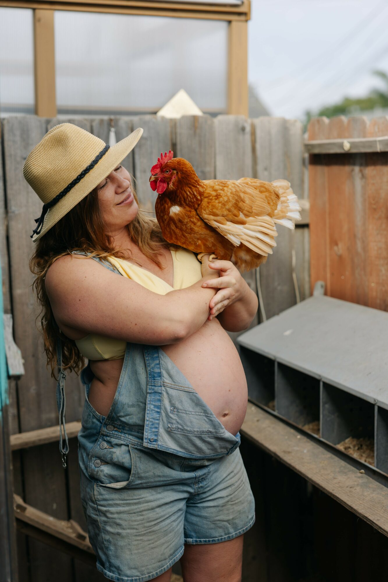 Pregnant woman holding a chicken outdoors, wearing a hat and casual clothes, in front of a wooden fence.