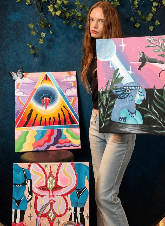 Young woman with long red hair holding three colorful paintings, standing against a dark background with green foliage.
