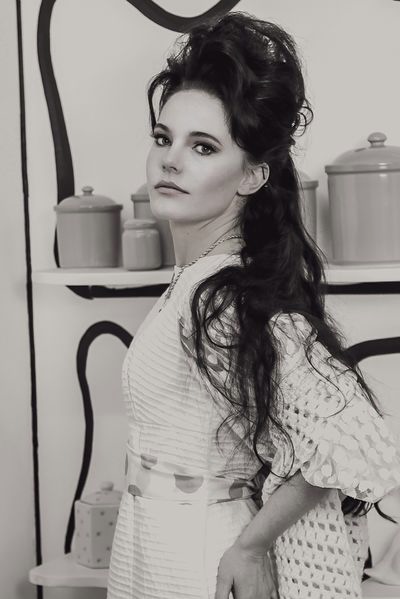 Woman with long dark hair and a patterned dress, standing indoors with shelves and containers behind her.