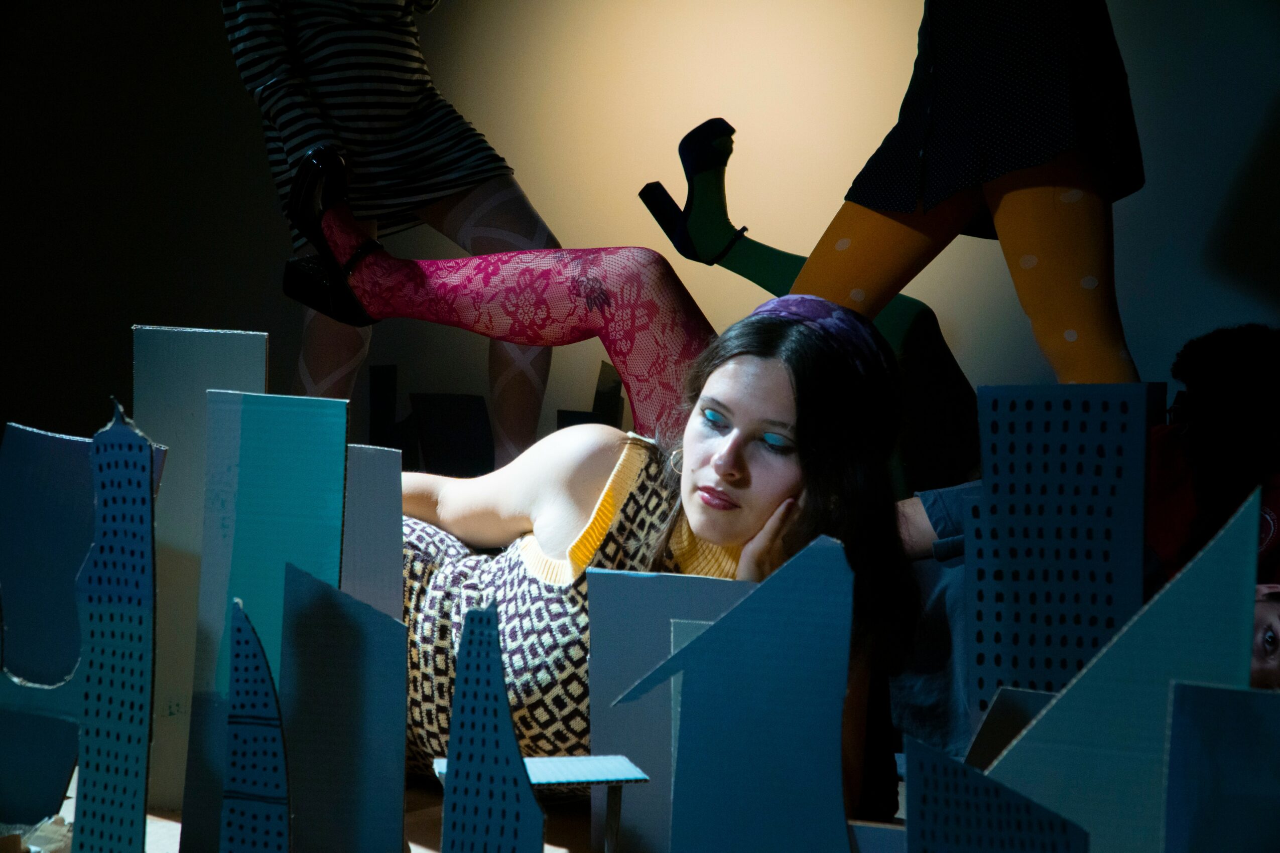 Woman lying on a table surrounded by blue geometric structures, with people in colorful clothing in the background.
