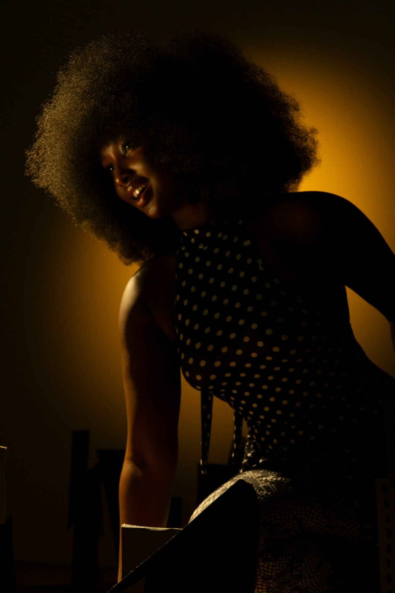 Person with curly hair smiling, wearing a polka dot top, illuminated by warm light, dark background.