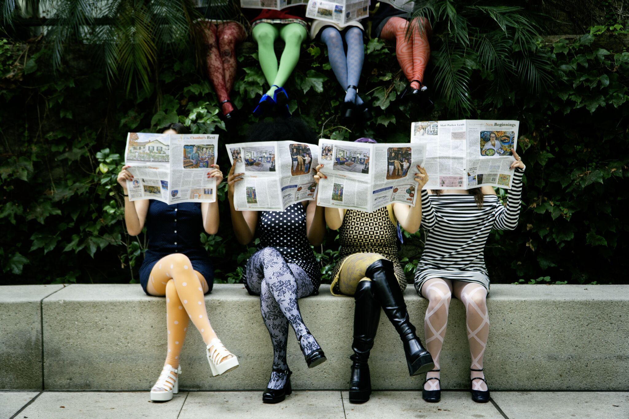 Four people sitting on a bench reading newspapers, with legs crossed and wearing different patterned outfits, in front of green foliage.