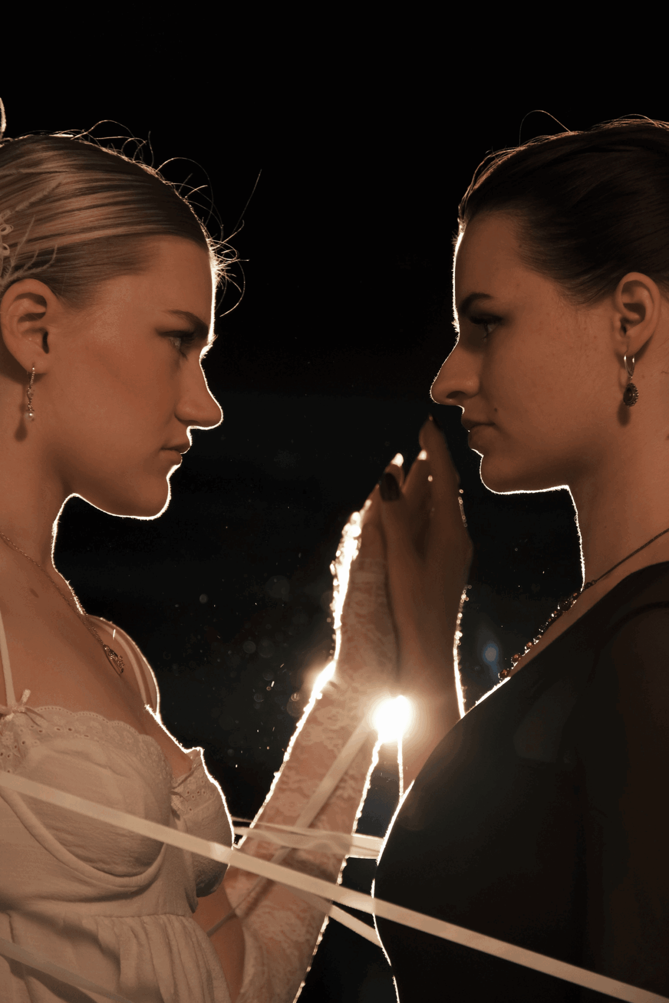 Two women face each other with hands touching, backlit by a bright light, dark background, close-up shot.