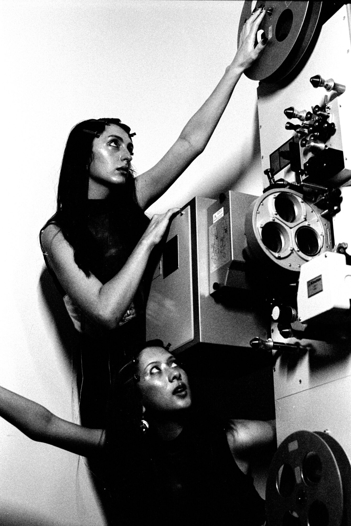 Two women operate vintage audio equipment, one reaching for a record on a wall-mounted turntable, in black and white.