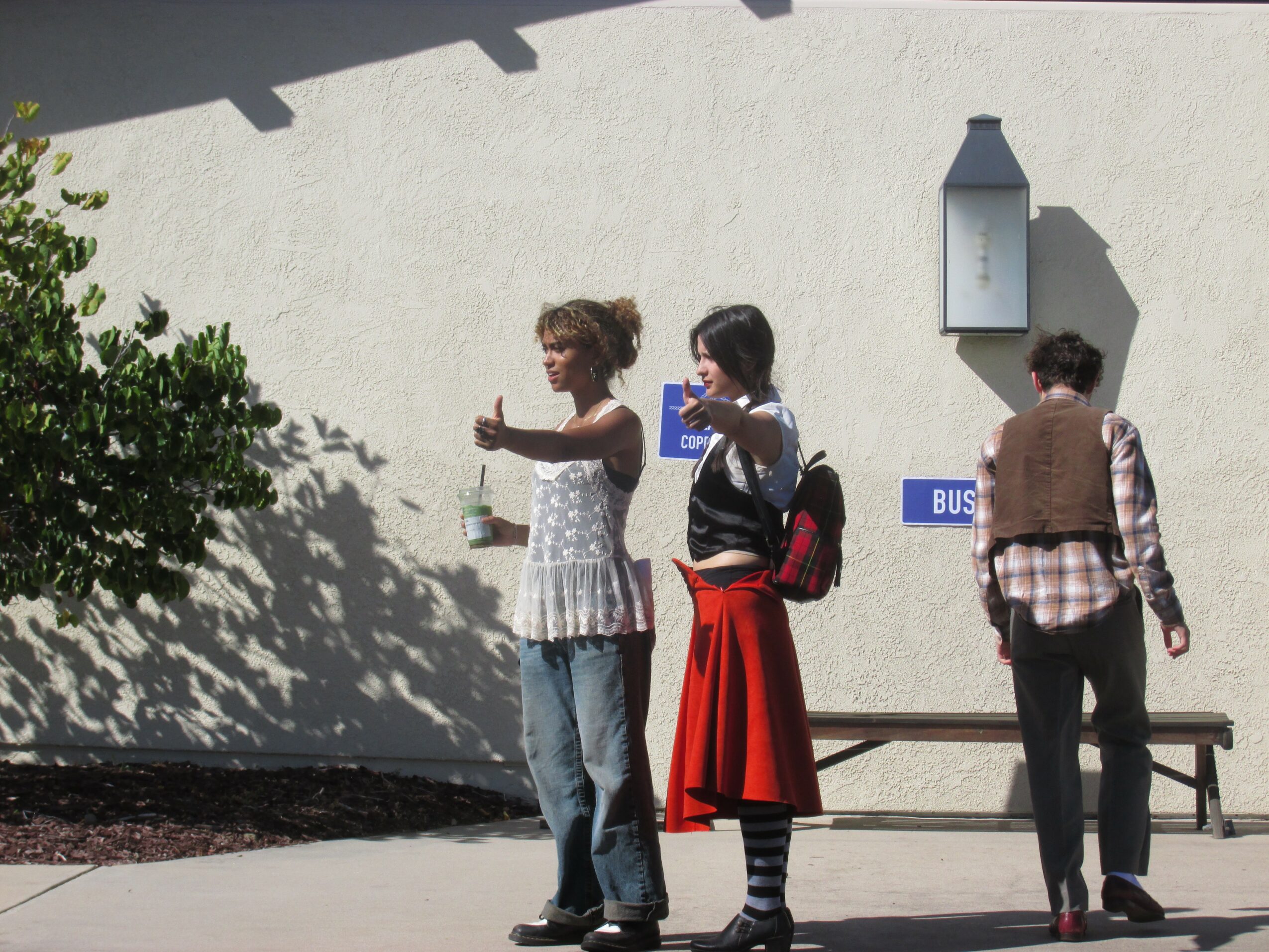 Three people waiting at a bus stop, two women and one man, standing near a white wall with a lamp and a bench.