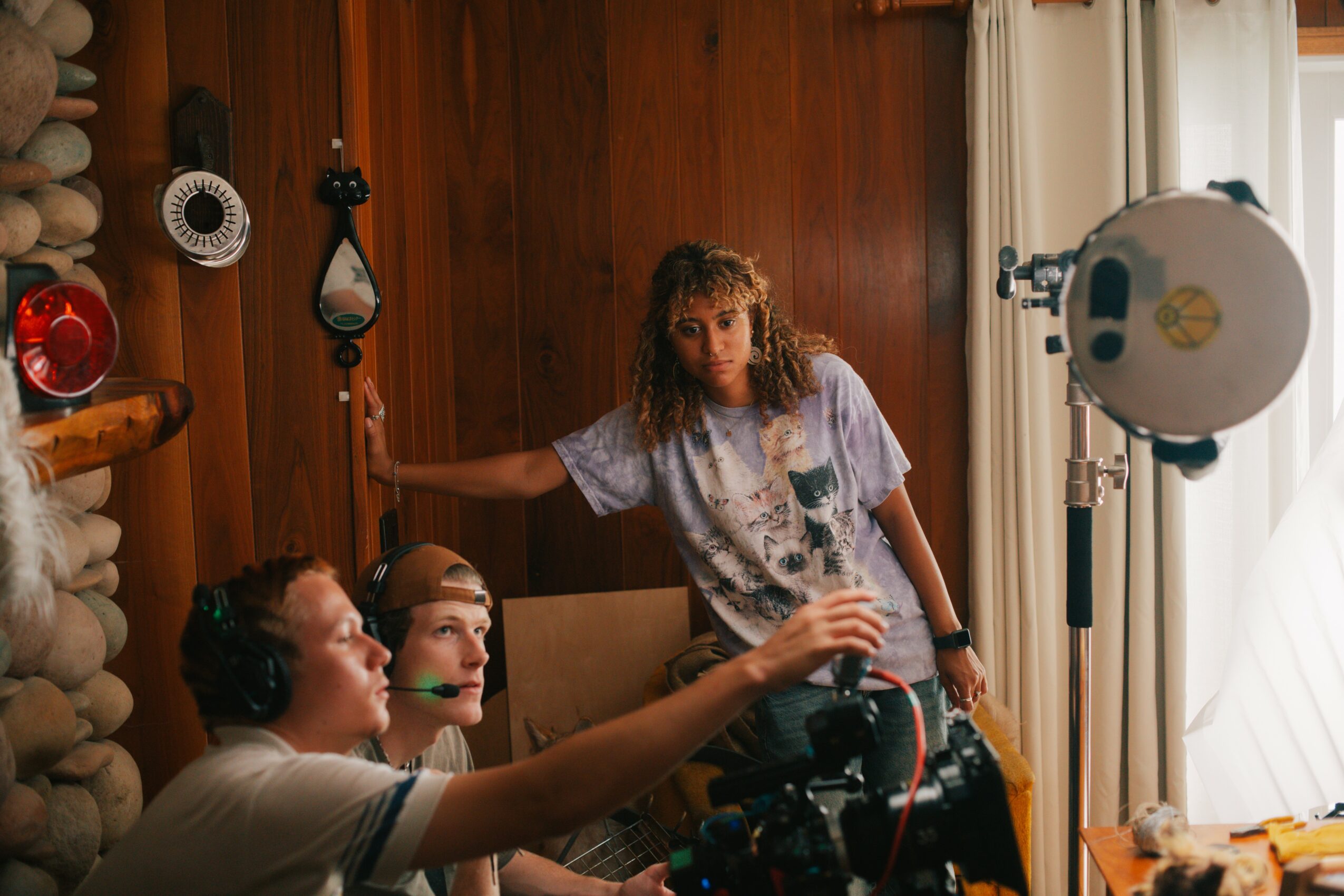 Three women in a room with wooden walls, one woman standing near a camera, two women sitting with headphones, one reaching for the camera.