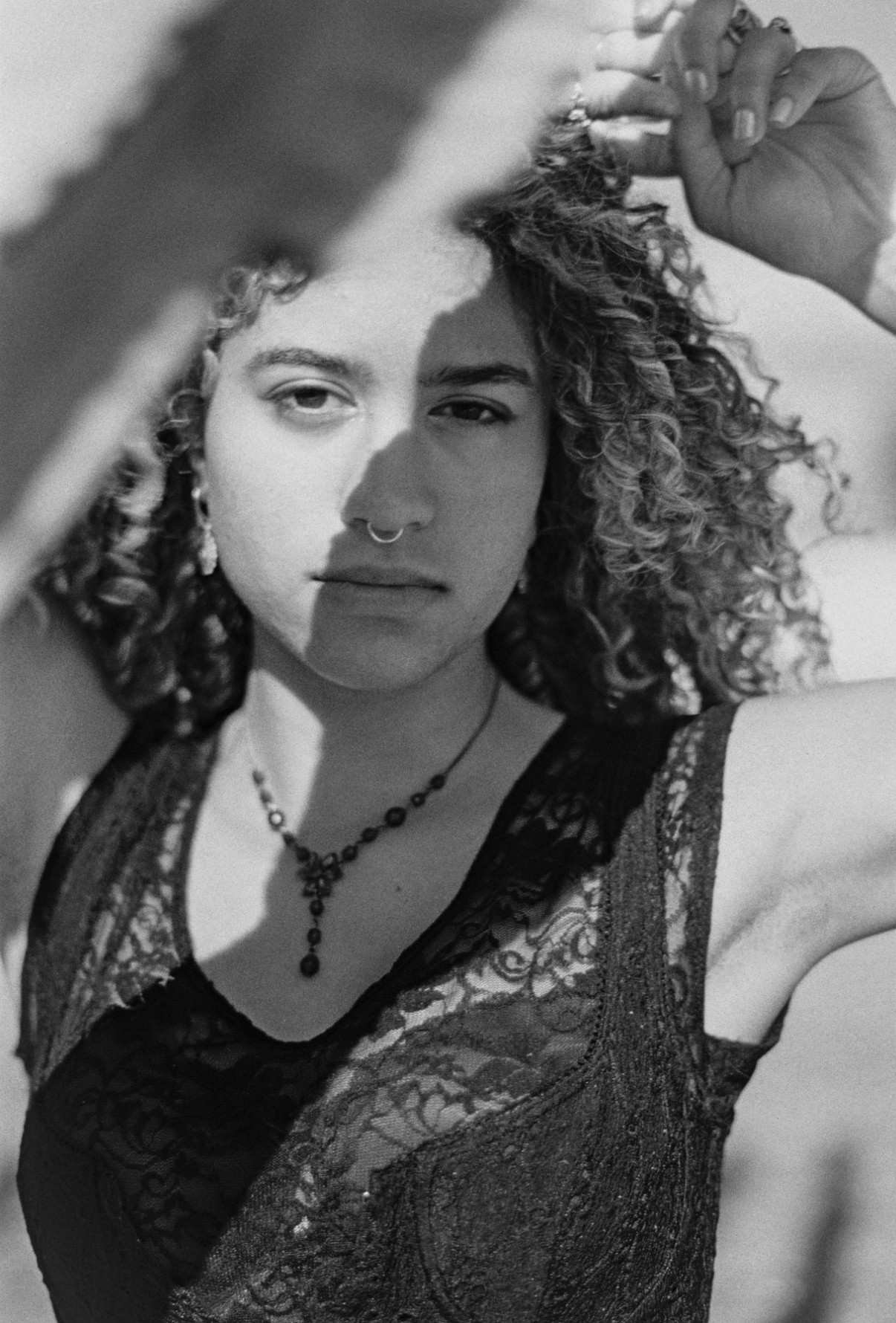 Woman with curly hair, wearing a lace top and necklace, looking at the camera with one arm raised.