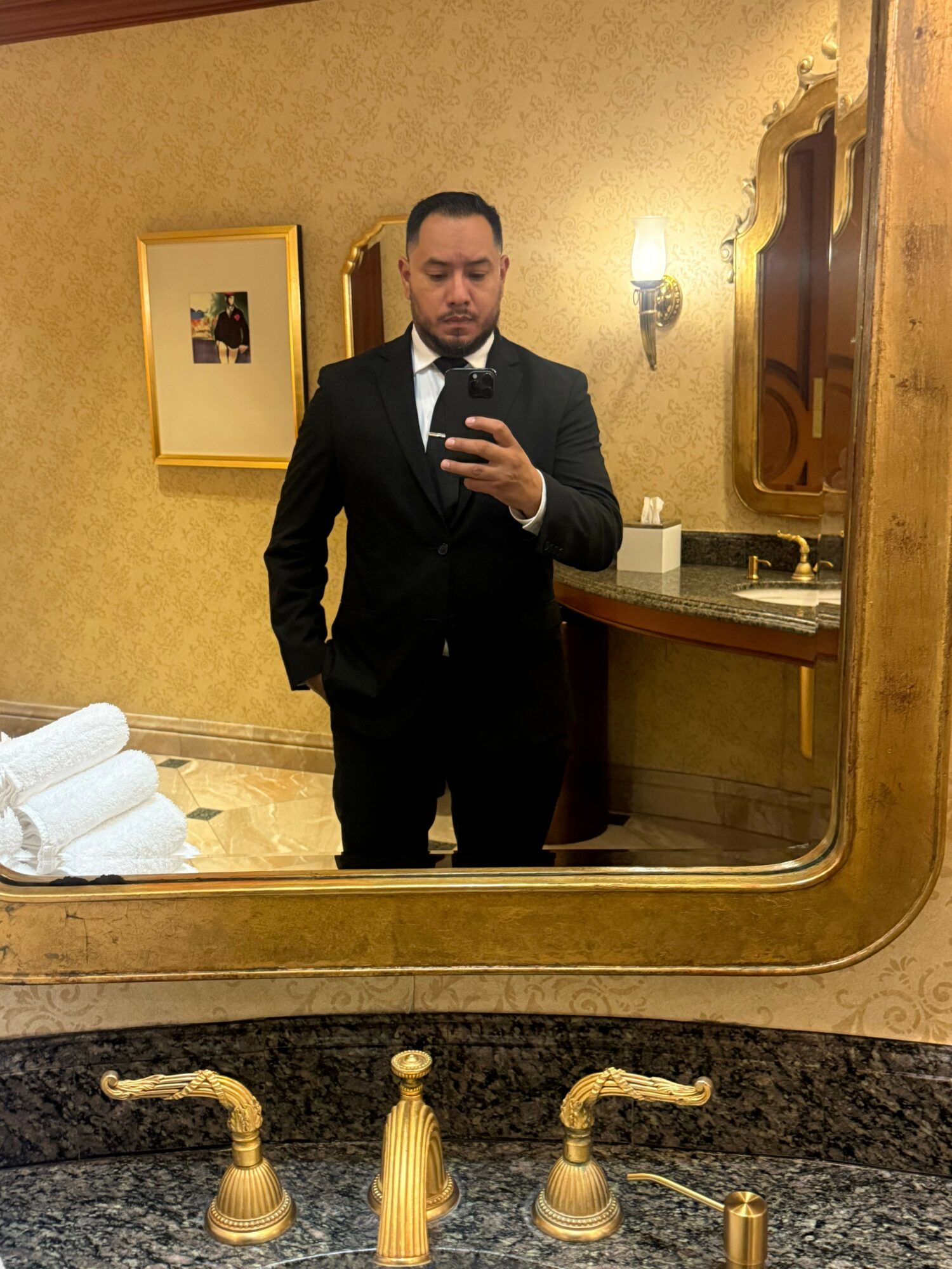 Man in a black suit taking a mirror selfie in a bathroom with gold fixtures and a framed picture on the wall.