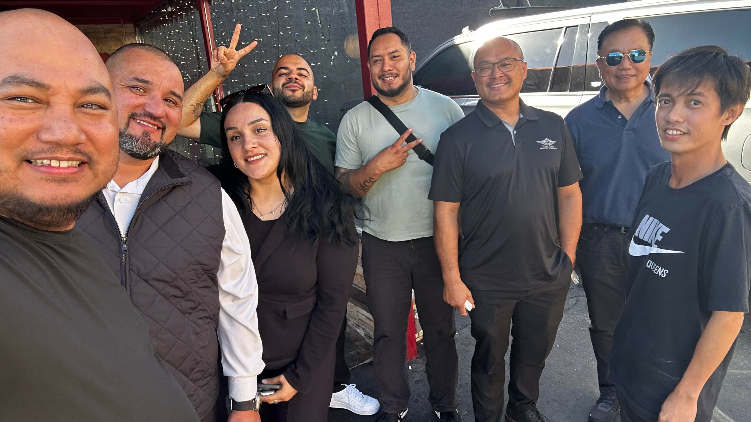 Group of nine people smiling and posing outdoors, some making peace signs, with a vehicle in the background.