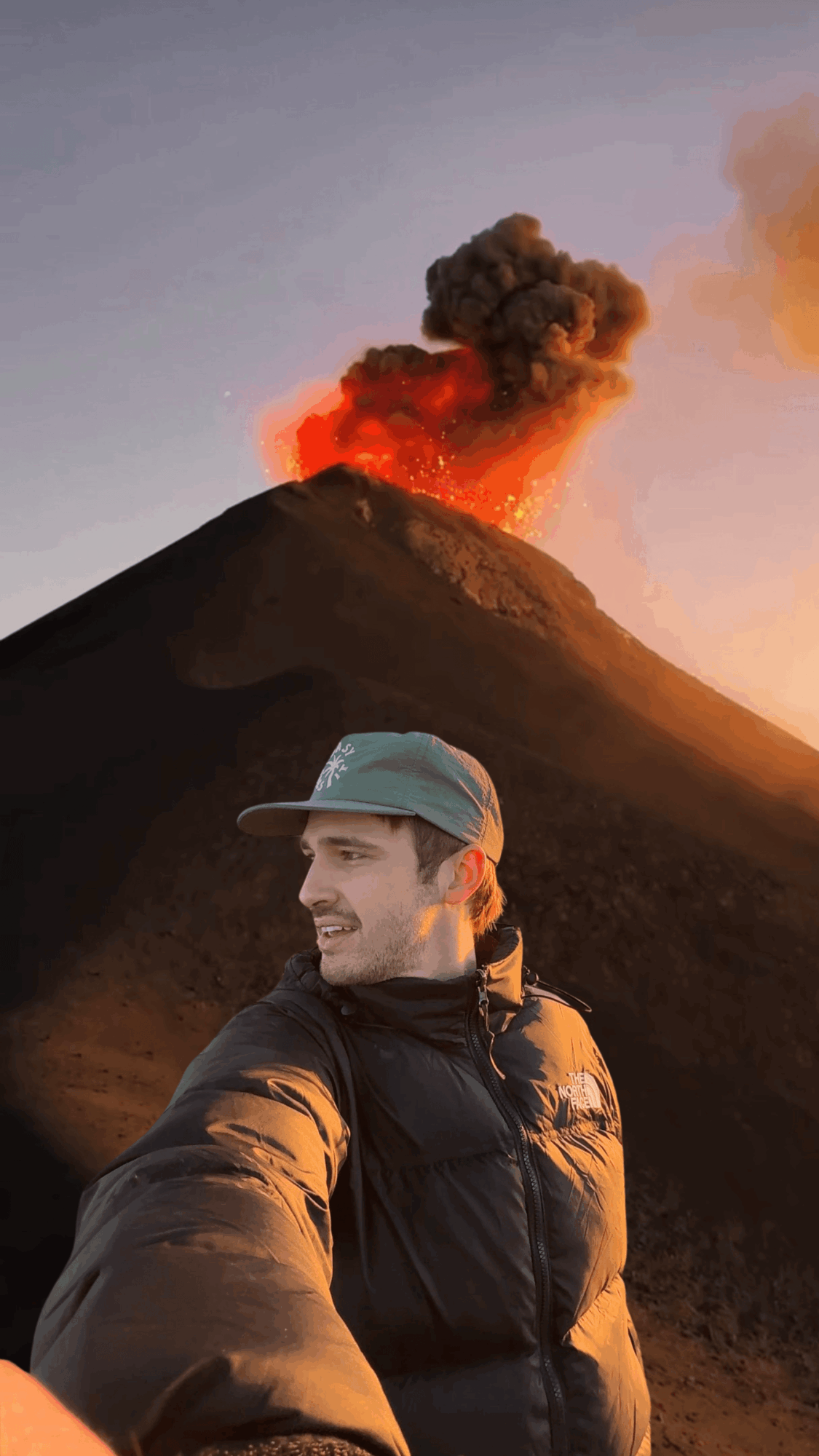 Young man in a cap and jacket taking a selfie with erupting volcano in background, ash and smoke rising into sky.
