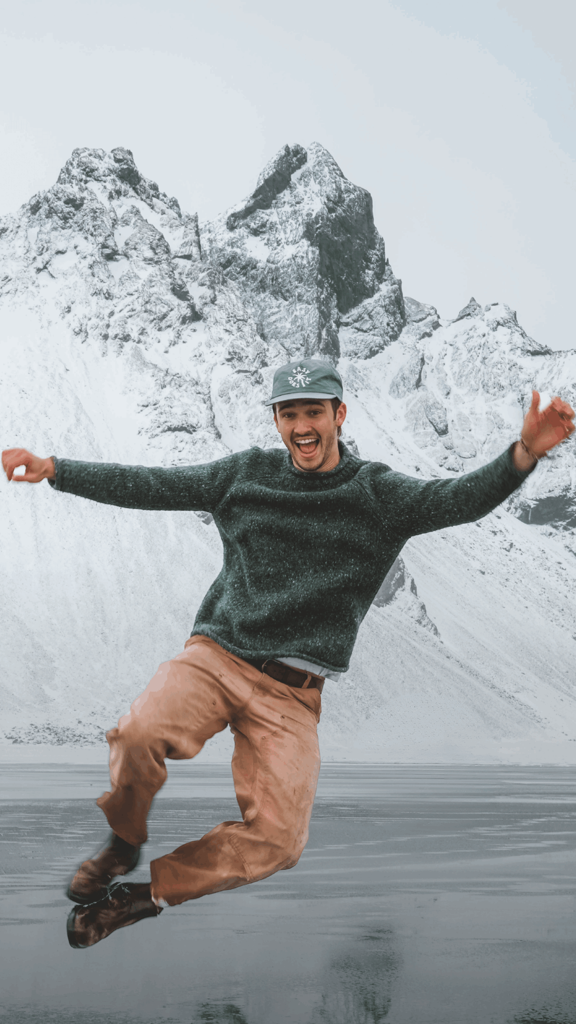 Person jumping in front of snow-covered mountains with arms outstretched, smiling, wearing a hat and winter clothing.
