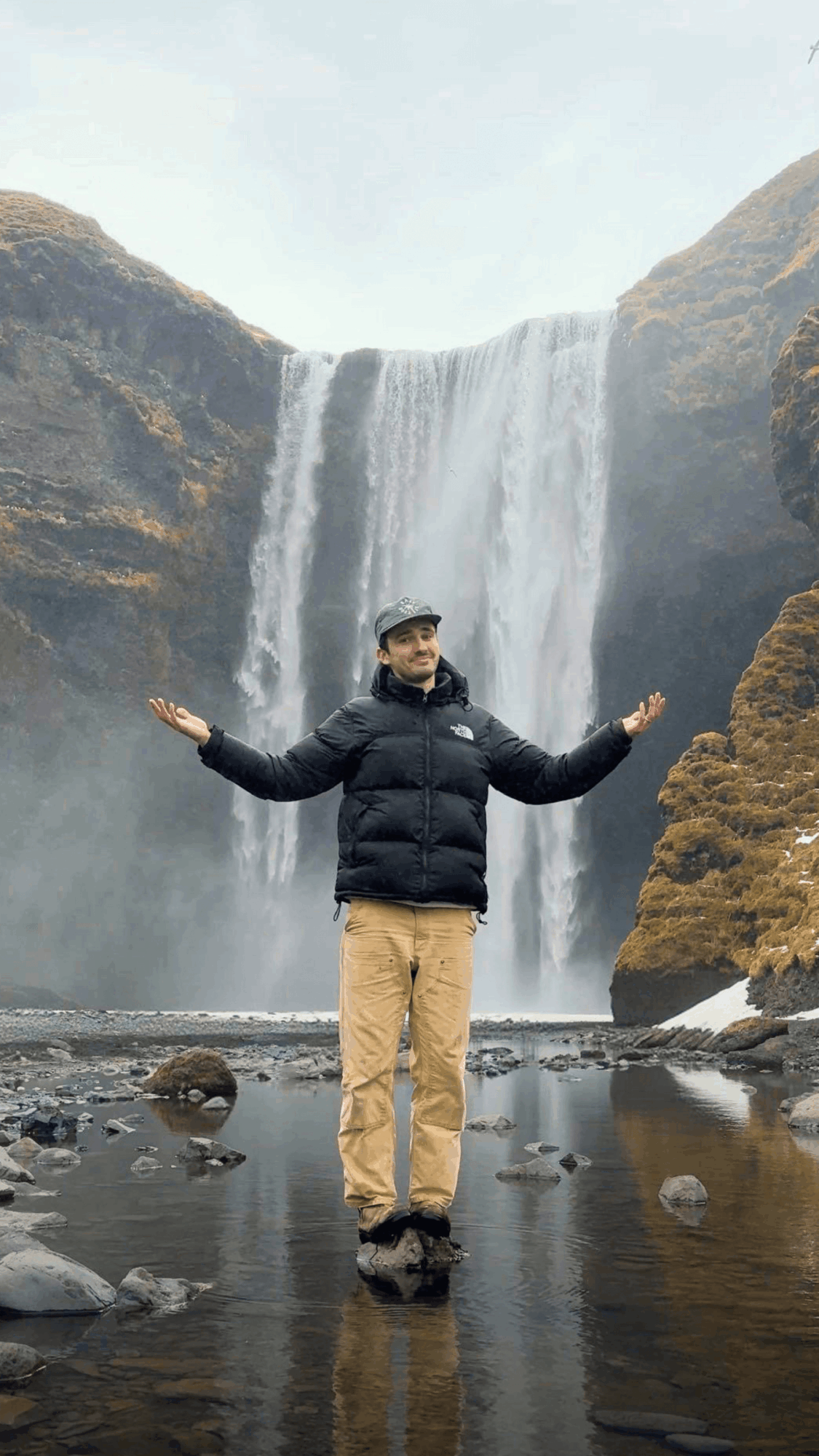 Person standing with arms outstretched in front of a waterfall, wearing a black jacket and beige pants.