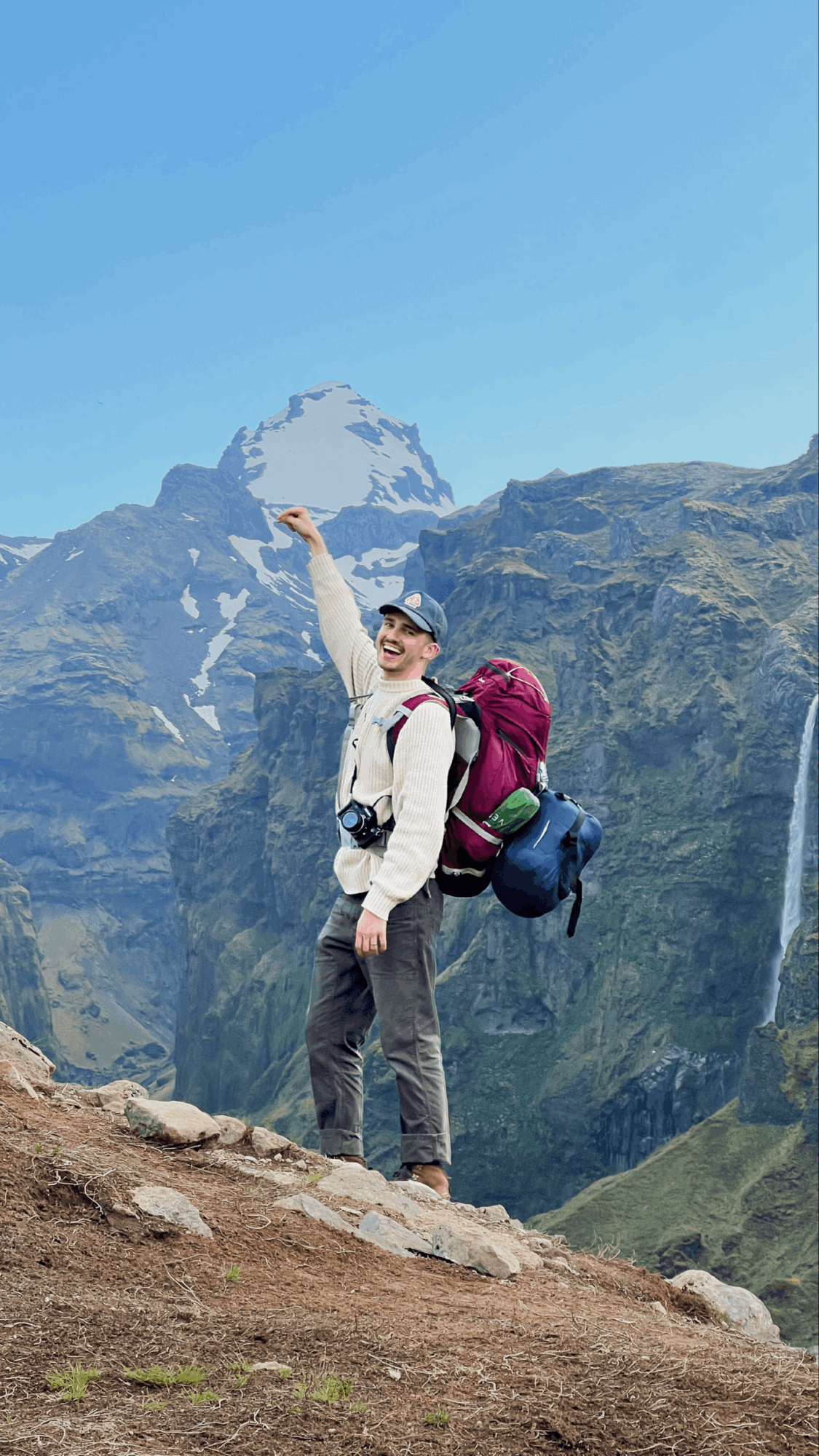 Person with a backpack standing on rocky terrain in front of mountains and waterfalls under a clear blue sky.