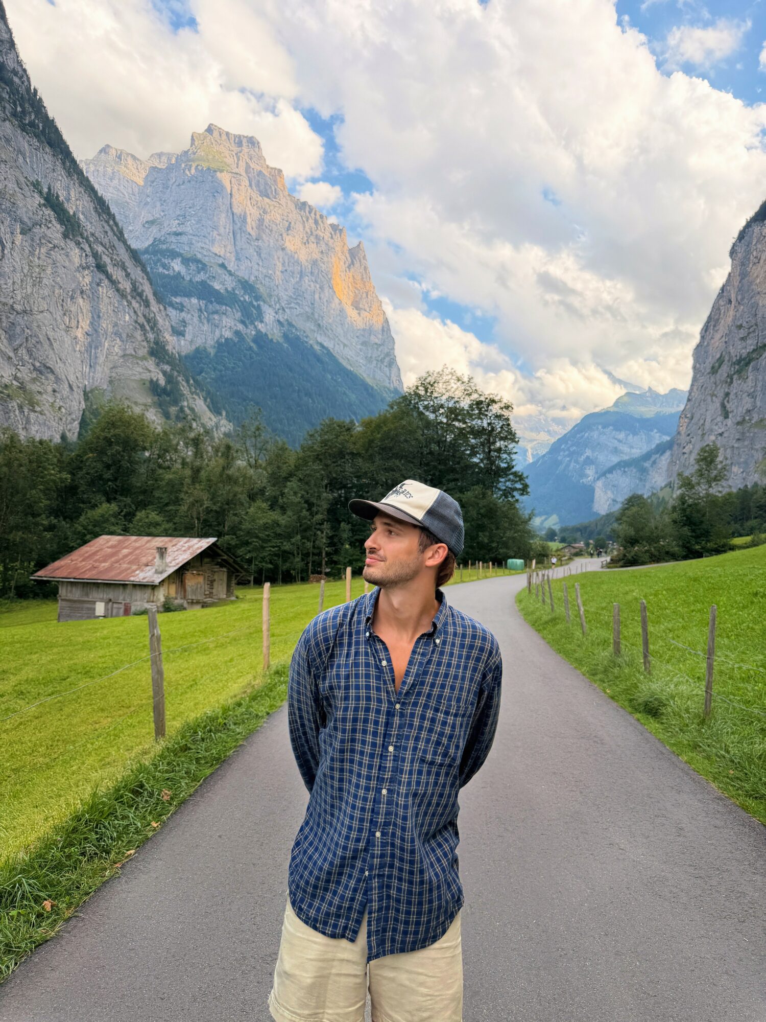 Young man in a checkered shirt and cap standing on a rural road with mountains and green fields in the background.