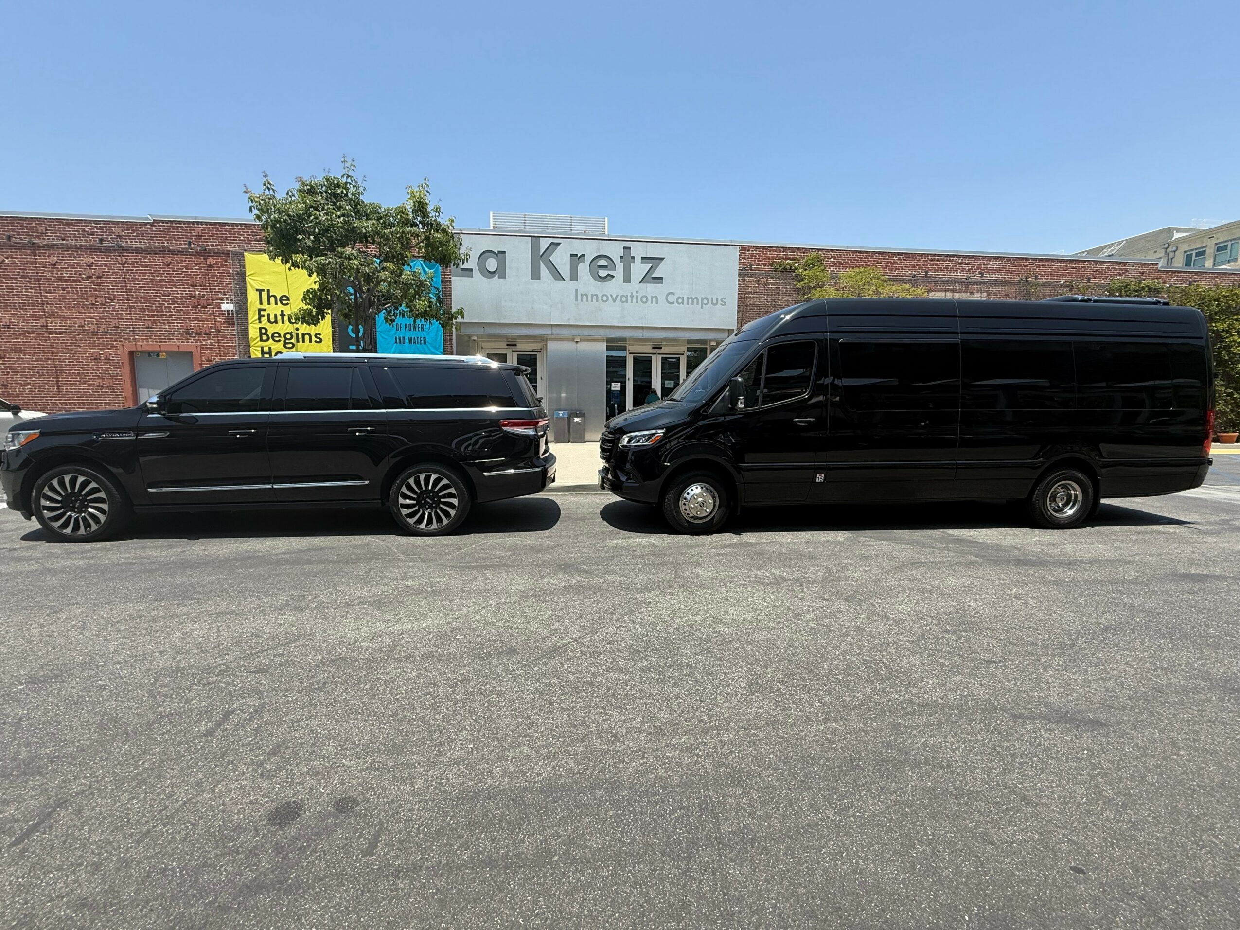 Two vehicles, a sedan and a van, parked side by side in front of a building with signs and a tree.
