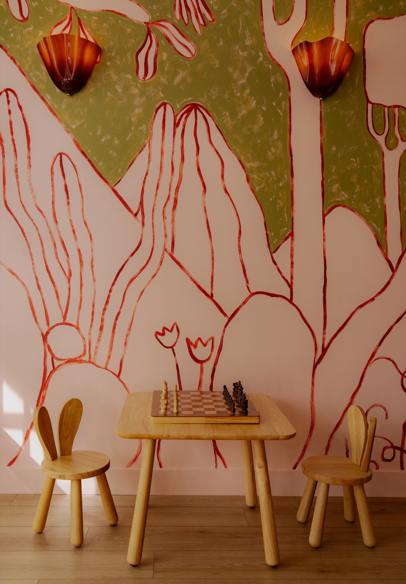 Children's play area with small chairs and a chessboard on a table, against a colorful mural of trees and mountains.