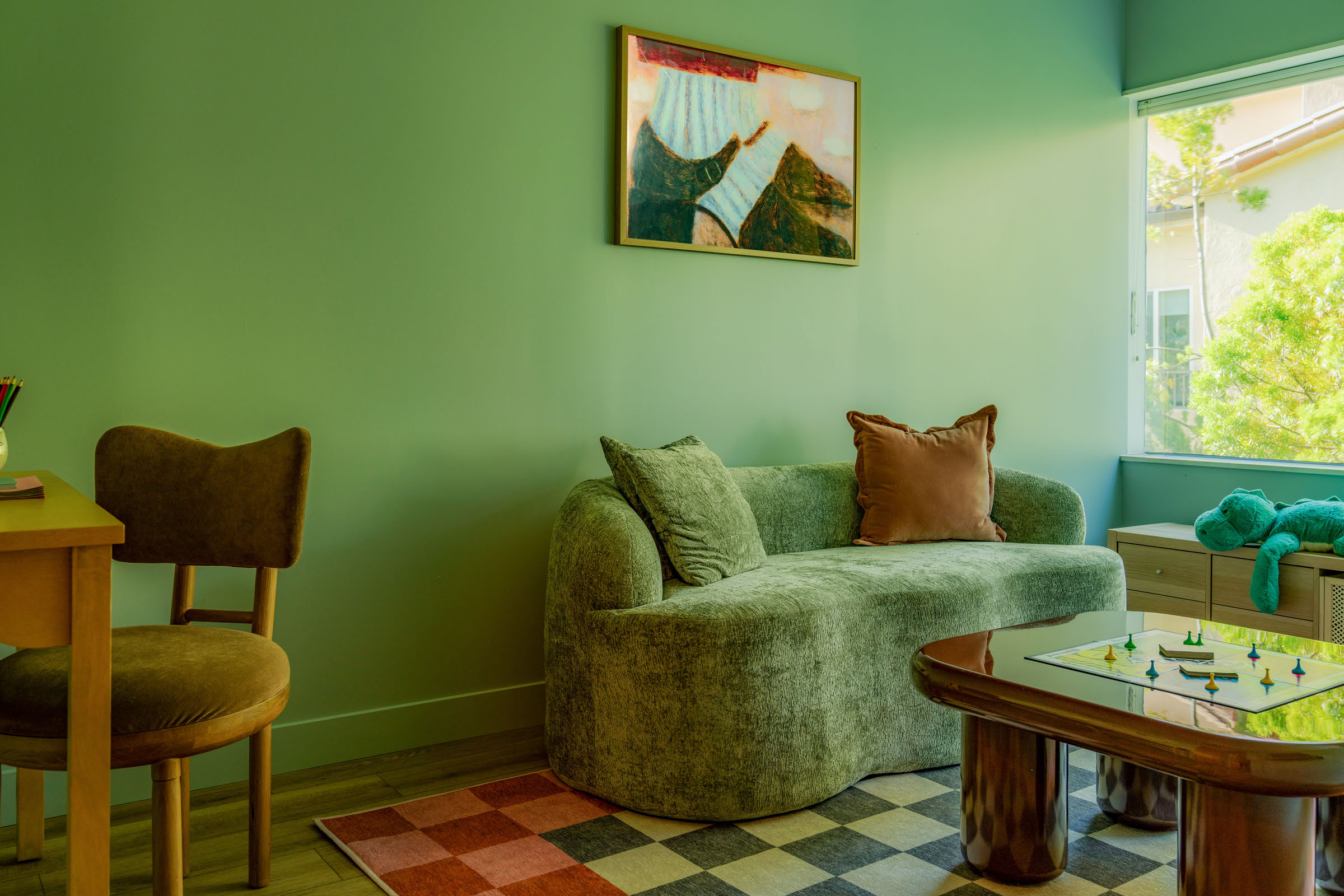 Living room with green walls, a green sofa with pillows, a wooden table, and a window with outdoor view.