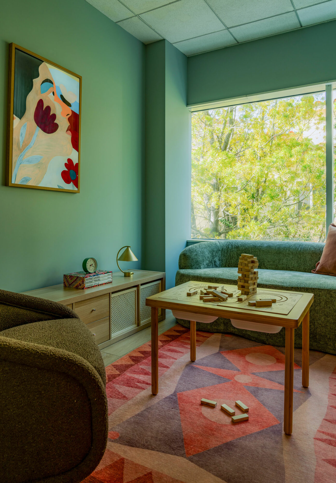Living room with green walls, a window with trees outside, a sofa, a coffee table with toys, and a colorful painting.