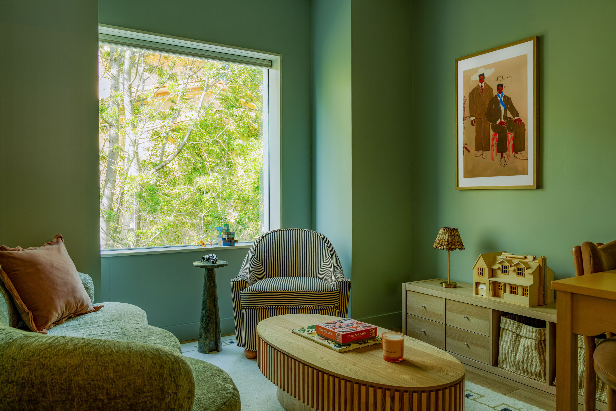 Living room with green walls, a window showing trees outside, a sofa, a chair, a coffee table, and framed artwork.