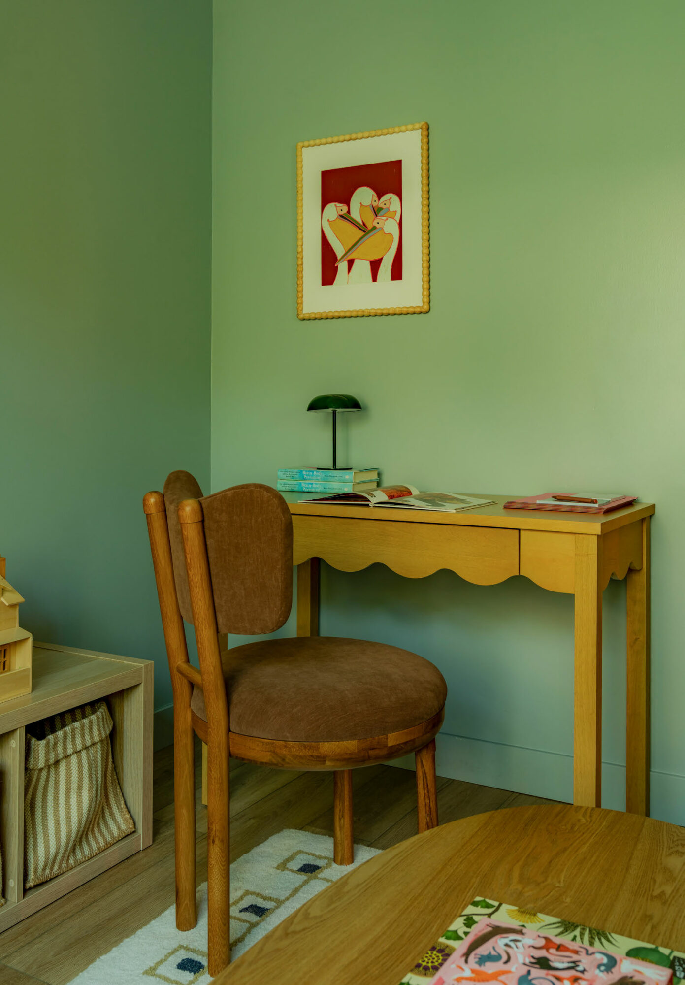 Corner of a room with a yellow desk, brown chair, framed artwork, and a small shelf with a basket.