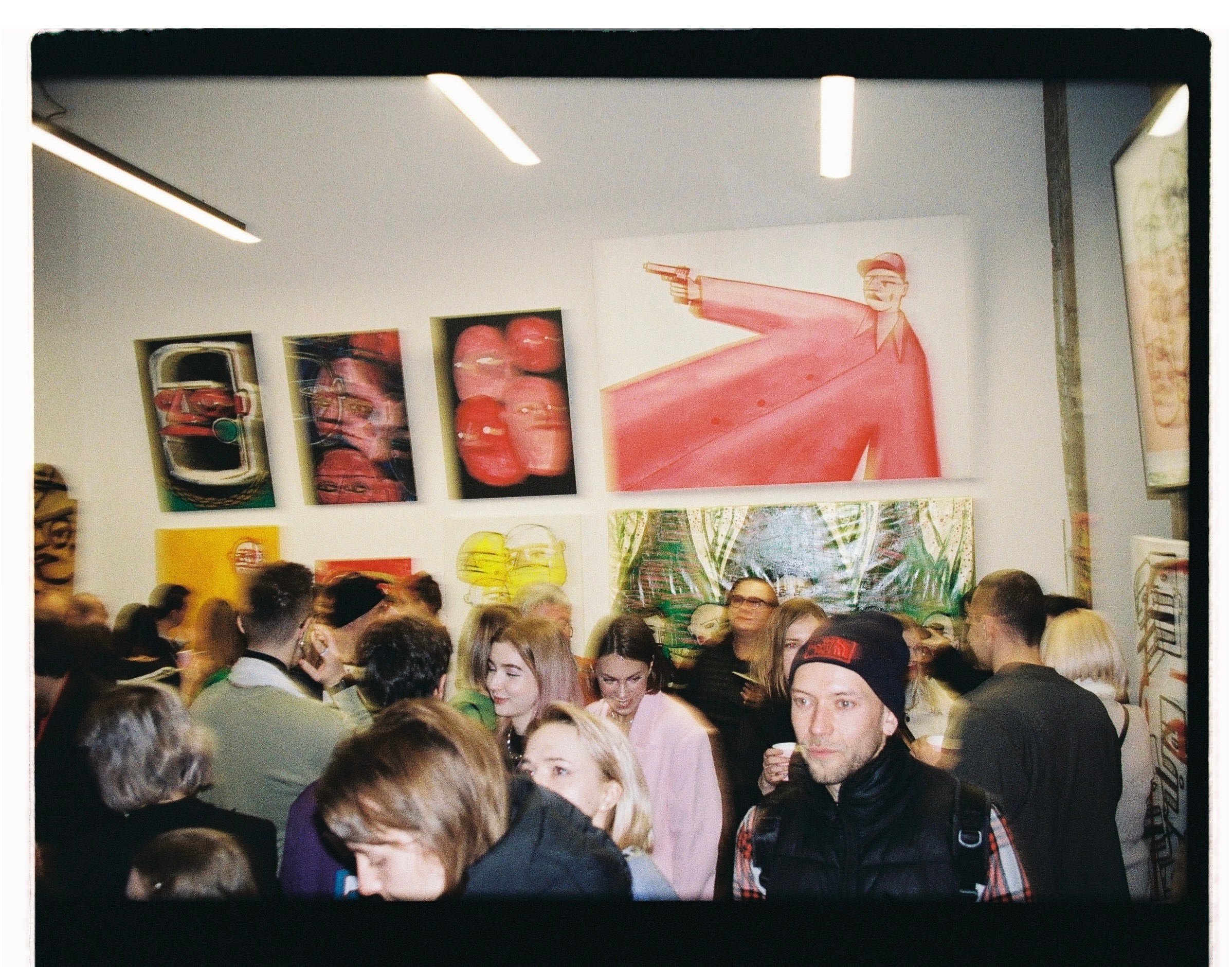 Crowd of people in an art gallery with colorful artwork on the walls, some looking at the art and others talking.