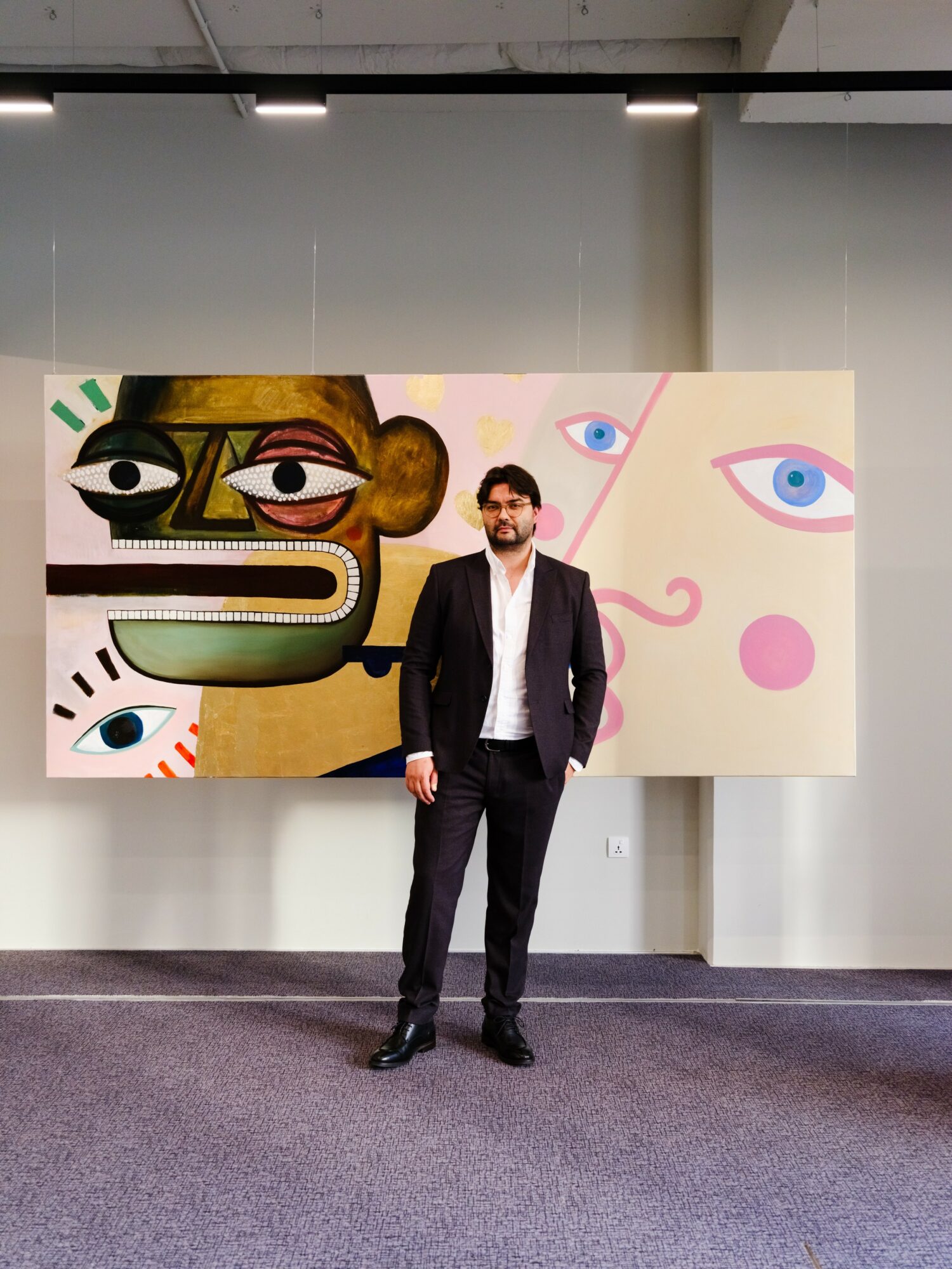 Man in a suit standing in front of colorful abstract artwork with eyes and faces, indoors.