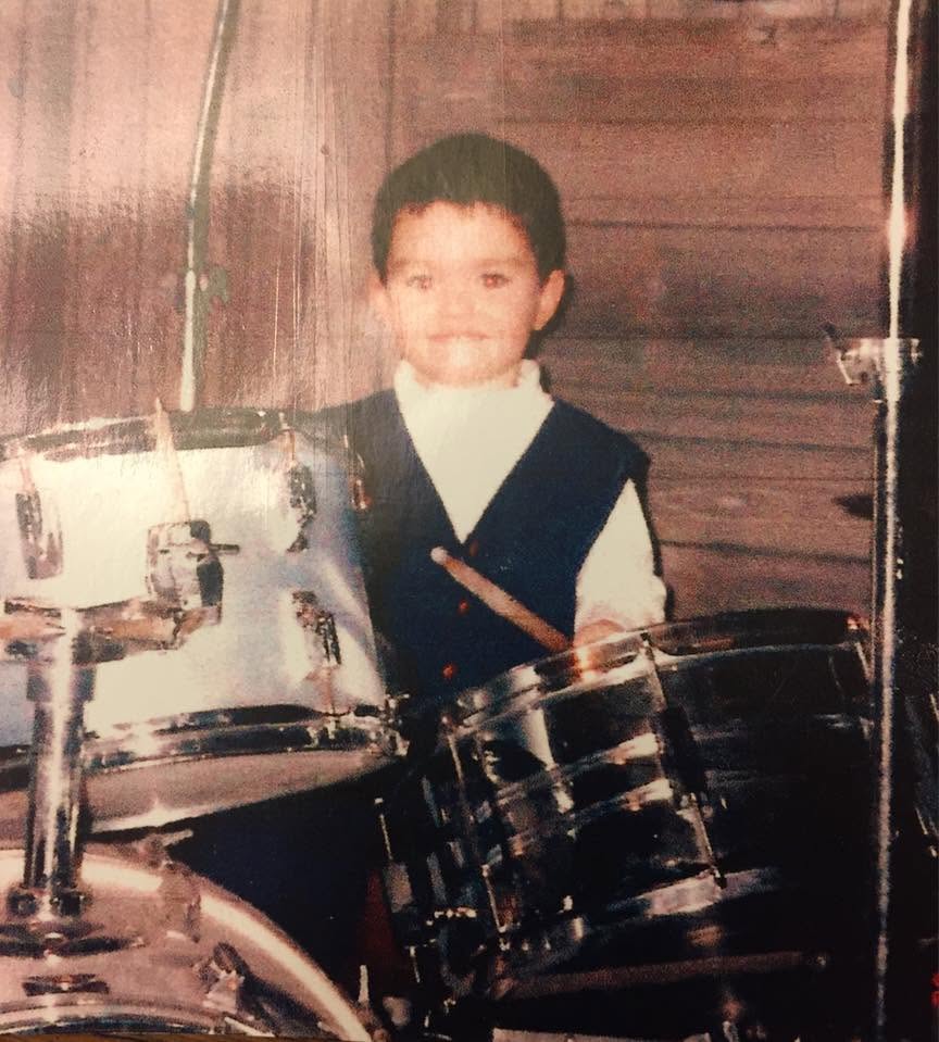 Child smiling, sitting behind a drum set, wearing a vest and white shirt, in front of wooden wall.