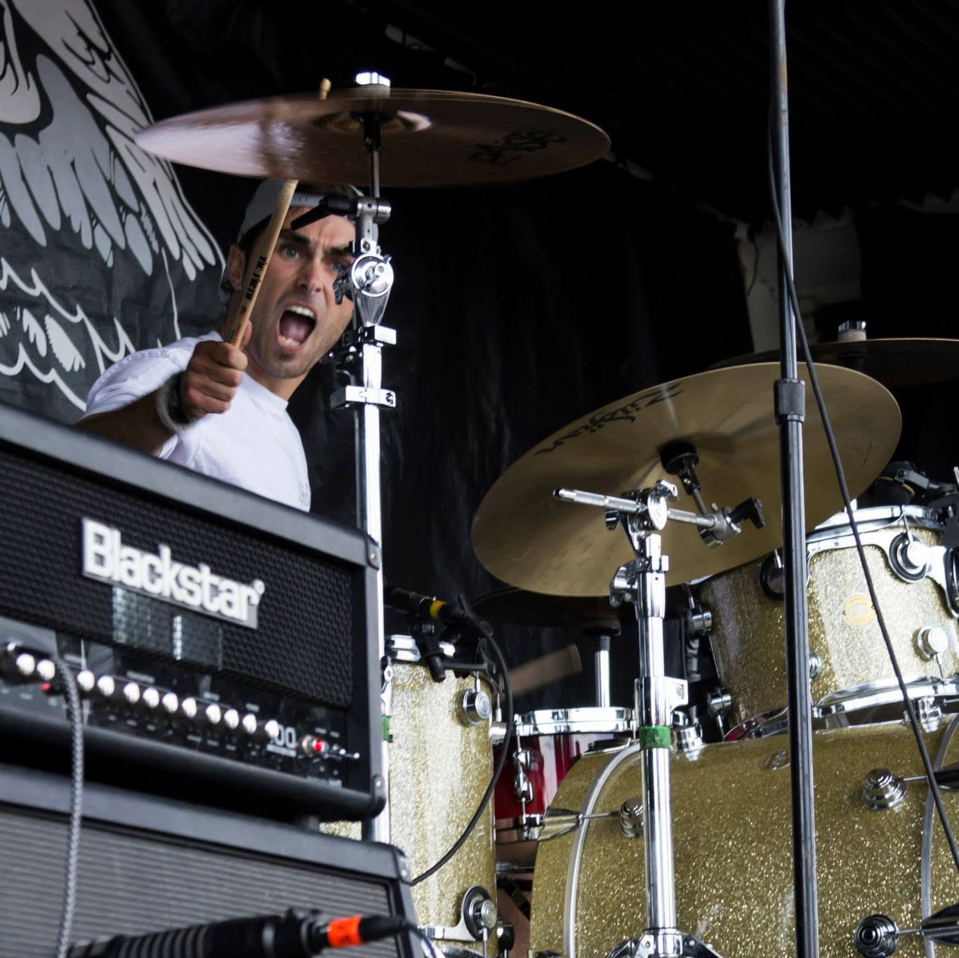 Musician playing drums and shouting, with drum set and Blackstar amplifier visible, on stage with dark background.